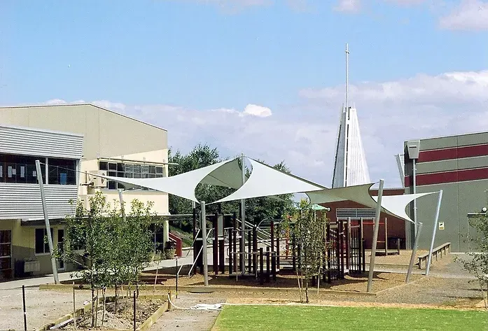 Custom shade sail canopy system covering outdoor amphitheatre at Beaconhills College in Berwick, featuring tensioned fabric in blue and beige tones over landscaped green play space with surrounding educational facilities.
