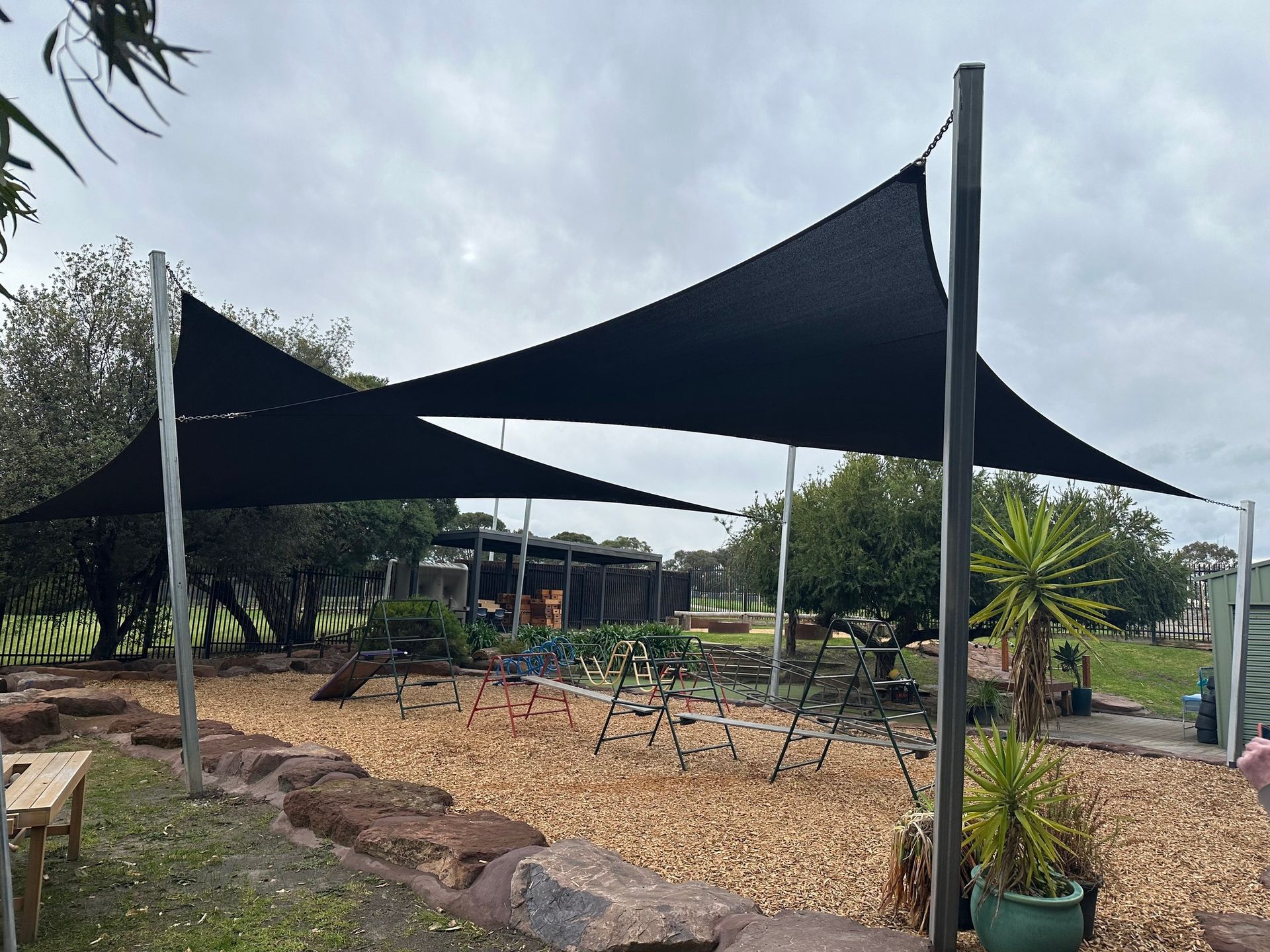 Black and blue hyperbolic commercial shade sails installed at Endeavourhills Kindergarten, providing effective sun protection over a timber-framed playground with swings, slide, and climbing equipment. Supported by tall steel posts and surrounded by natural landscaping including rocks, trees, and mulch ground cover. Custom-designed by a licensed shade sail installer in Melbourne