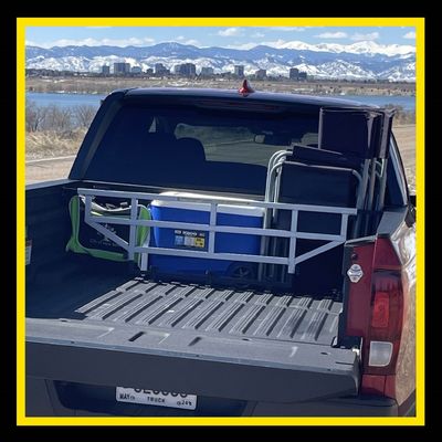Truck bed with cargo. White metal frame, blue cooler, chairs, and gear. Background has water and mountains.
