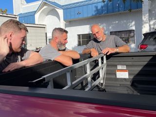Three men examining something in the back of a red pickup truck. Blue building in background.