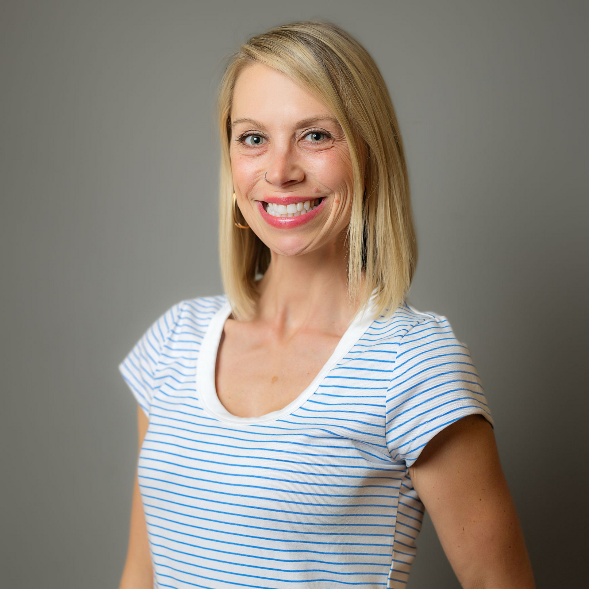 A smiling person with blonde shoulder-length hair wearing a blue and white striped t-shirt against a gray background.