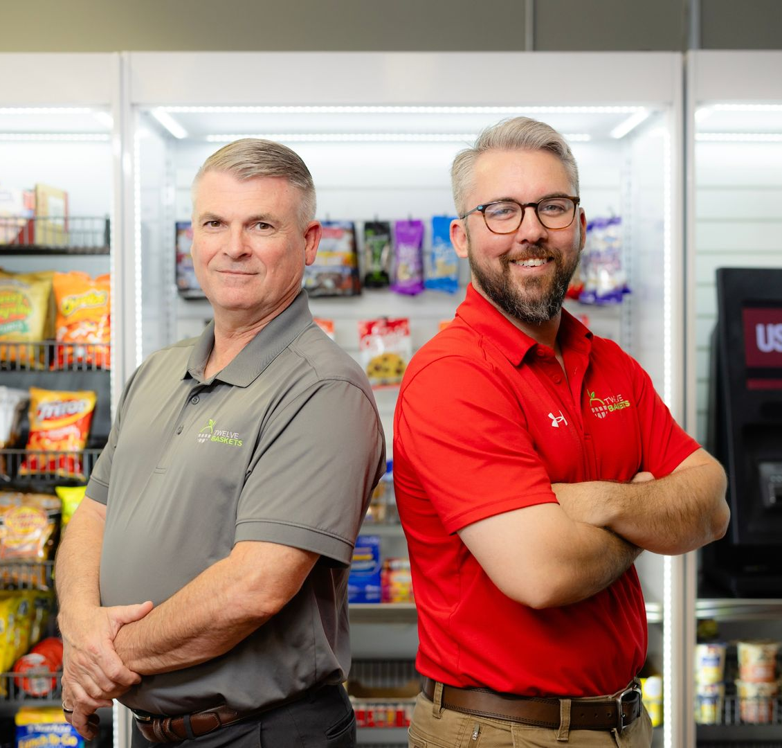 Two men in polo shirts stand in front of a well-stocked micro market display with snack shelves and a self-checkout kiosk.