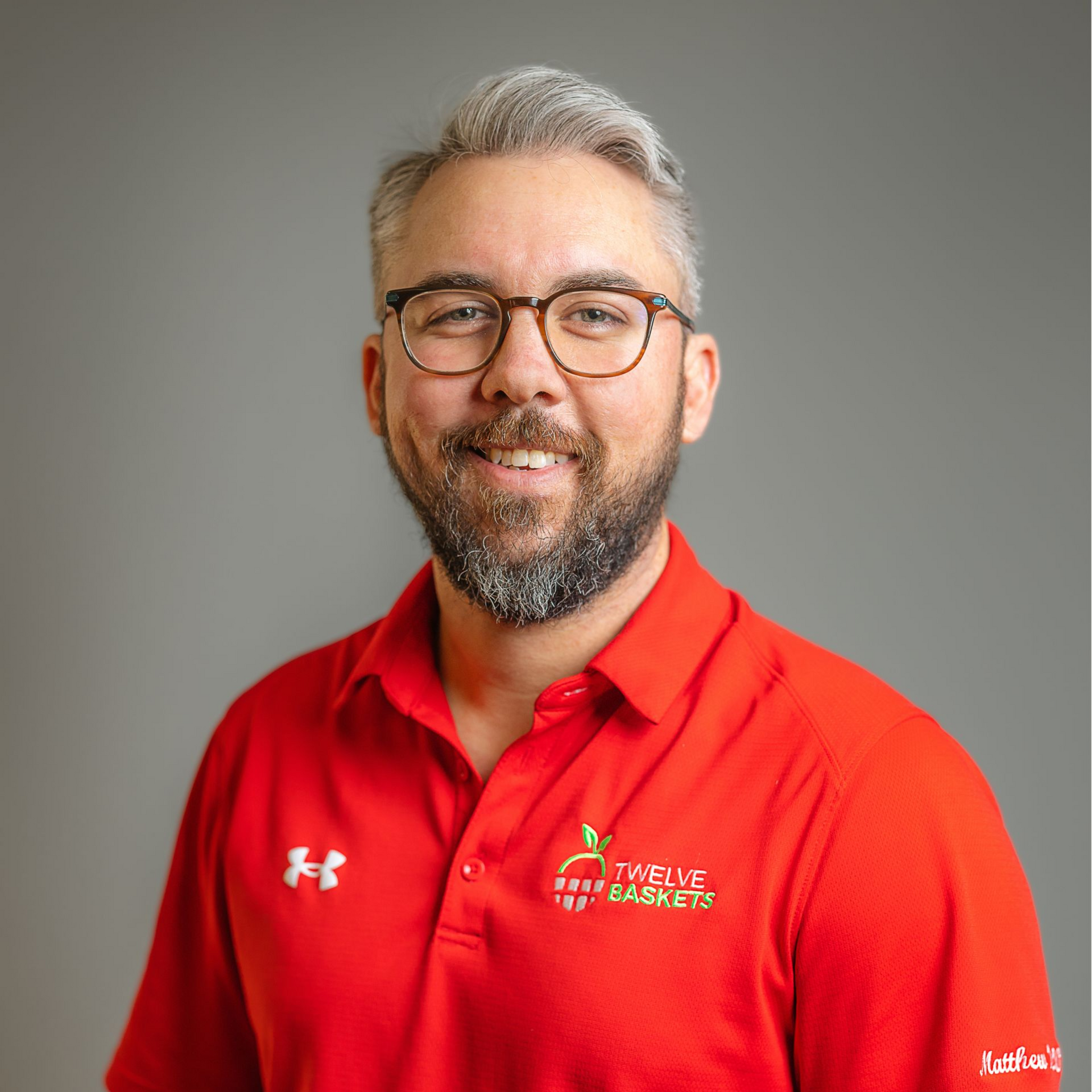 A person with a beard and glasses smiles, wearing a red short-sleeved polo shirt against a plain grey background.