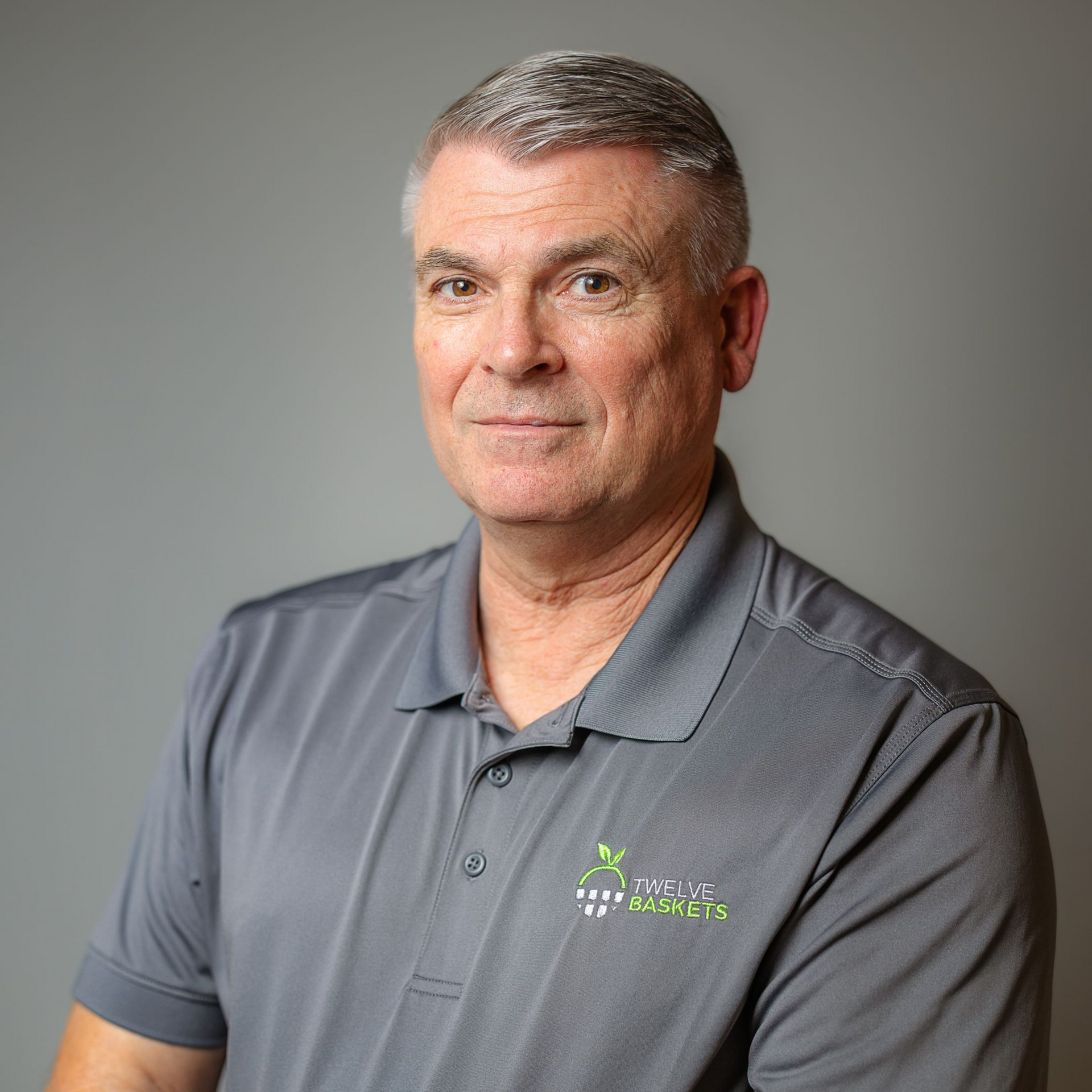 A person with short, graying hair wearing a gray polo shirt with a small green logo, posed against a gray background.