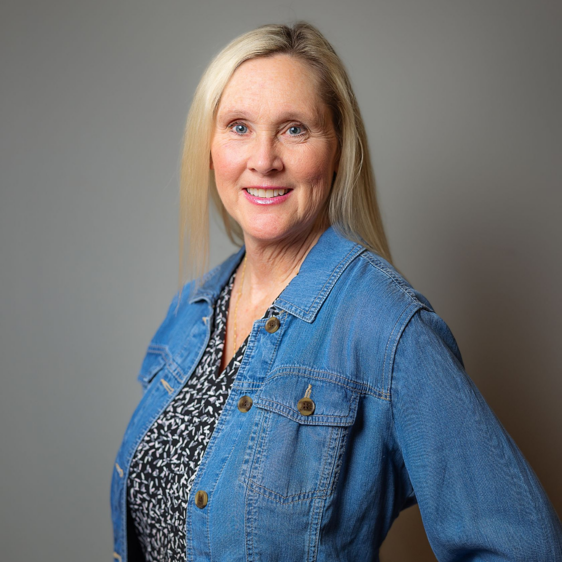 A woman with blonde hair, wearing a patterned top and a denim jacket, smiles while posing against a plain gray background.