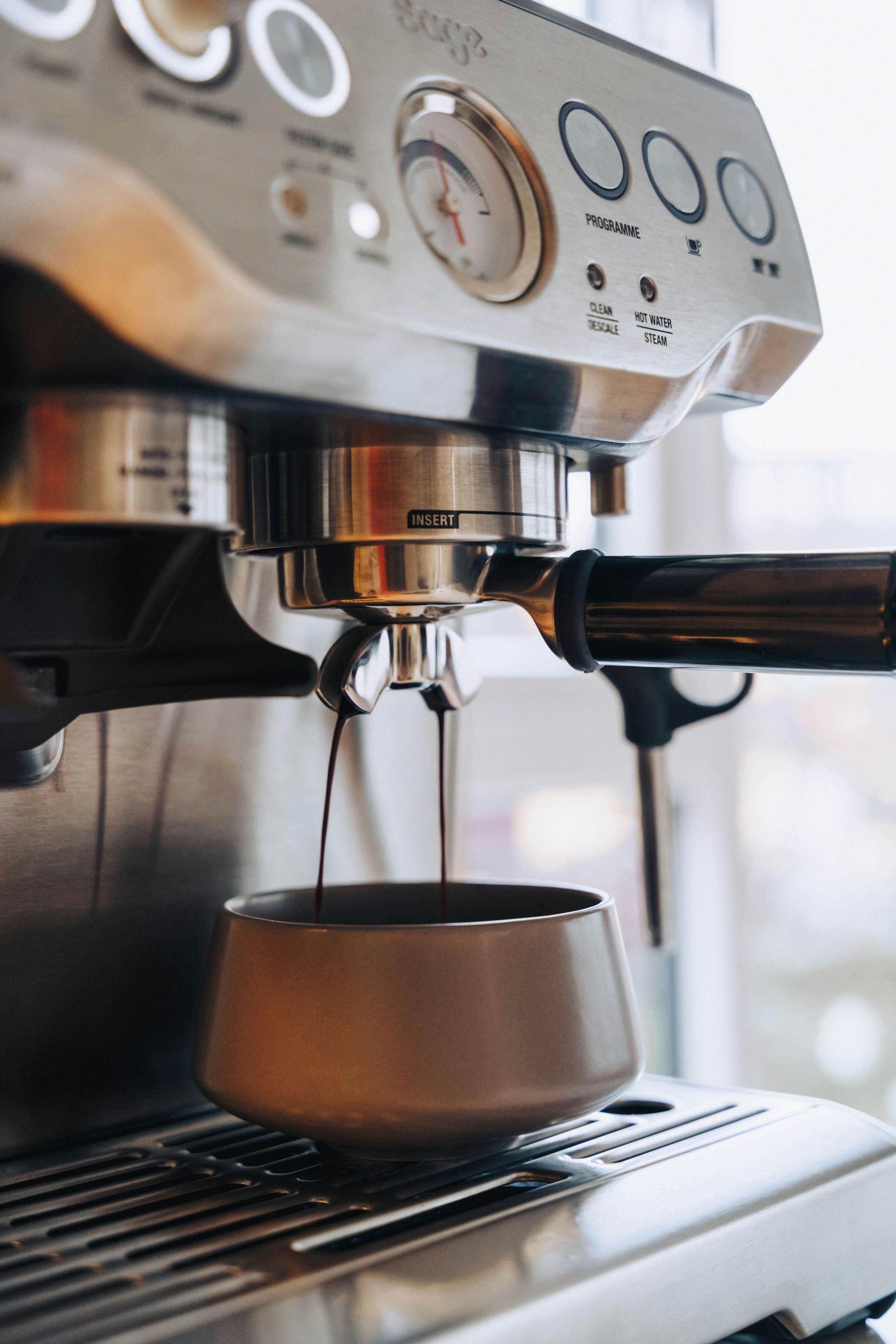 An espresso machine pours two streams of dark coffee into a brown ceramic cup resting on a stainless steel tray.