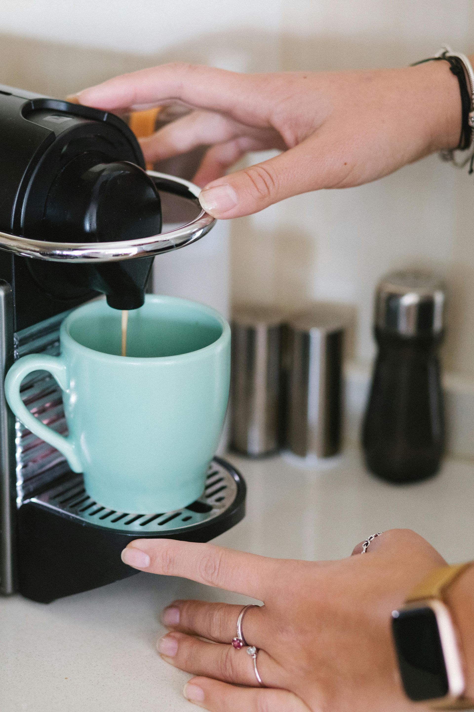 A person’s hand operates a black espresso machine, brewing coffee into a light blue mug on a white countertop.