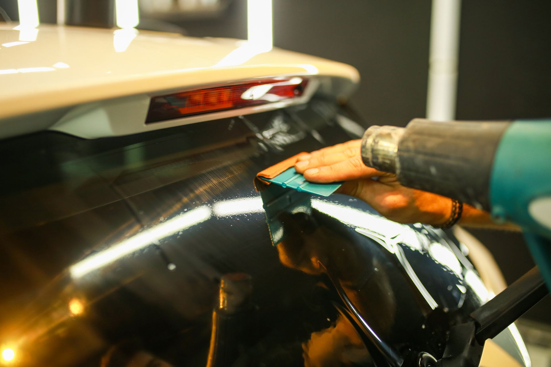 Person applying tint to a car window, using a heat gun and smoothing tool in a garage.