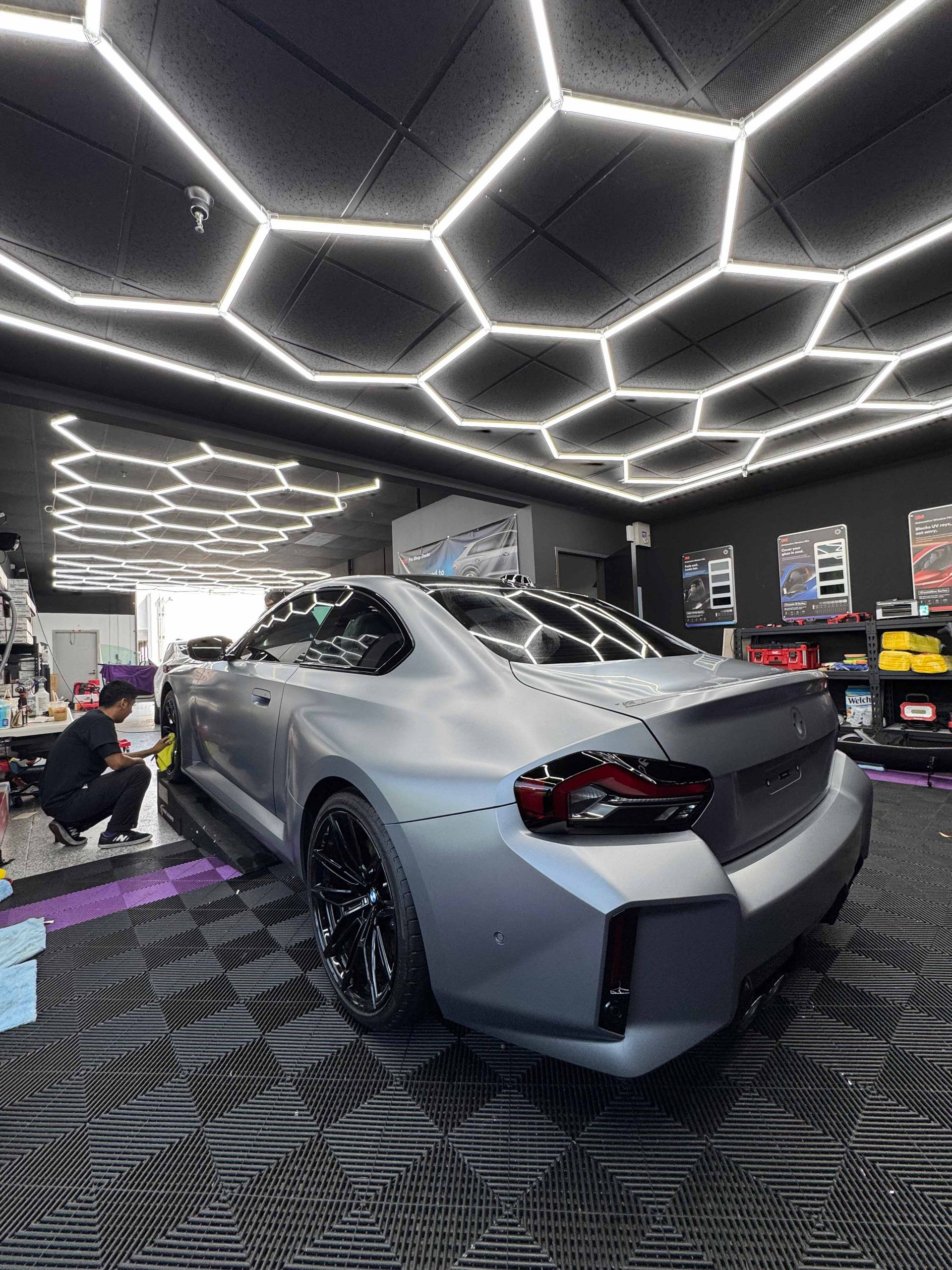 Gray car in a shop with honeycomb lights, a worker details the side.