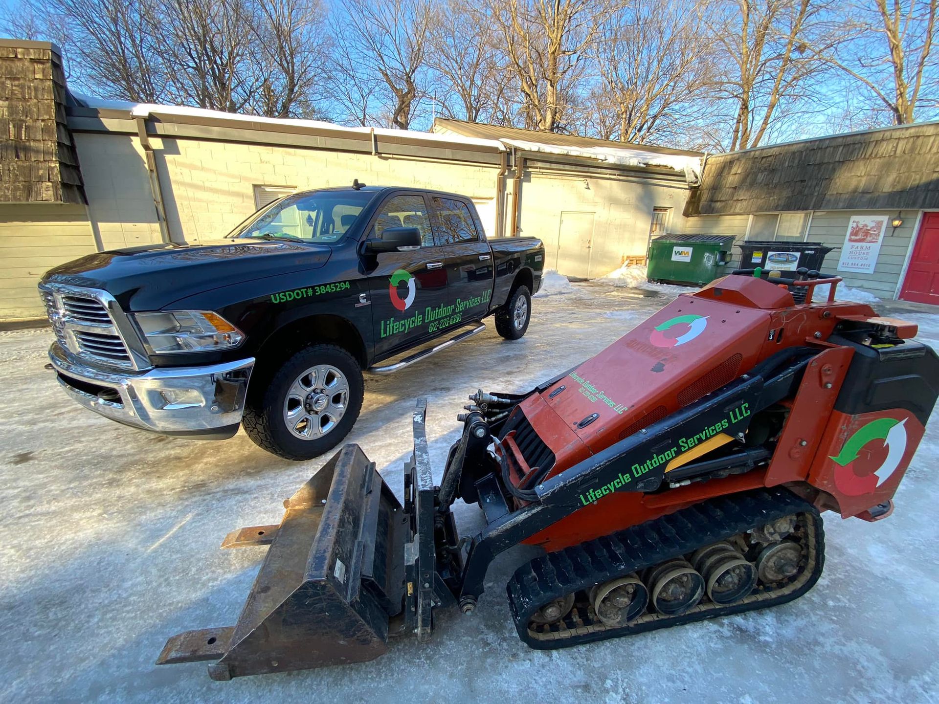 Black pickup truck and red skid steer loader parked in a snowy area. Both have company logos.