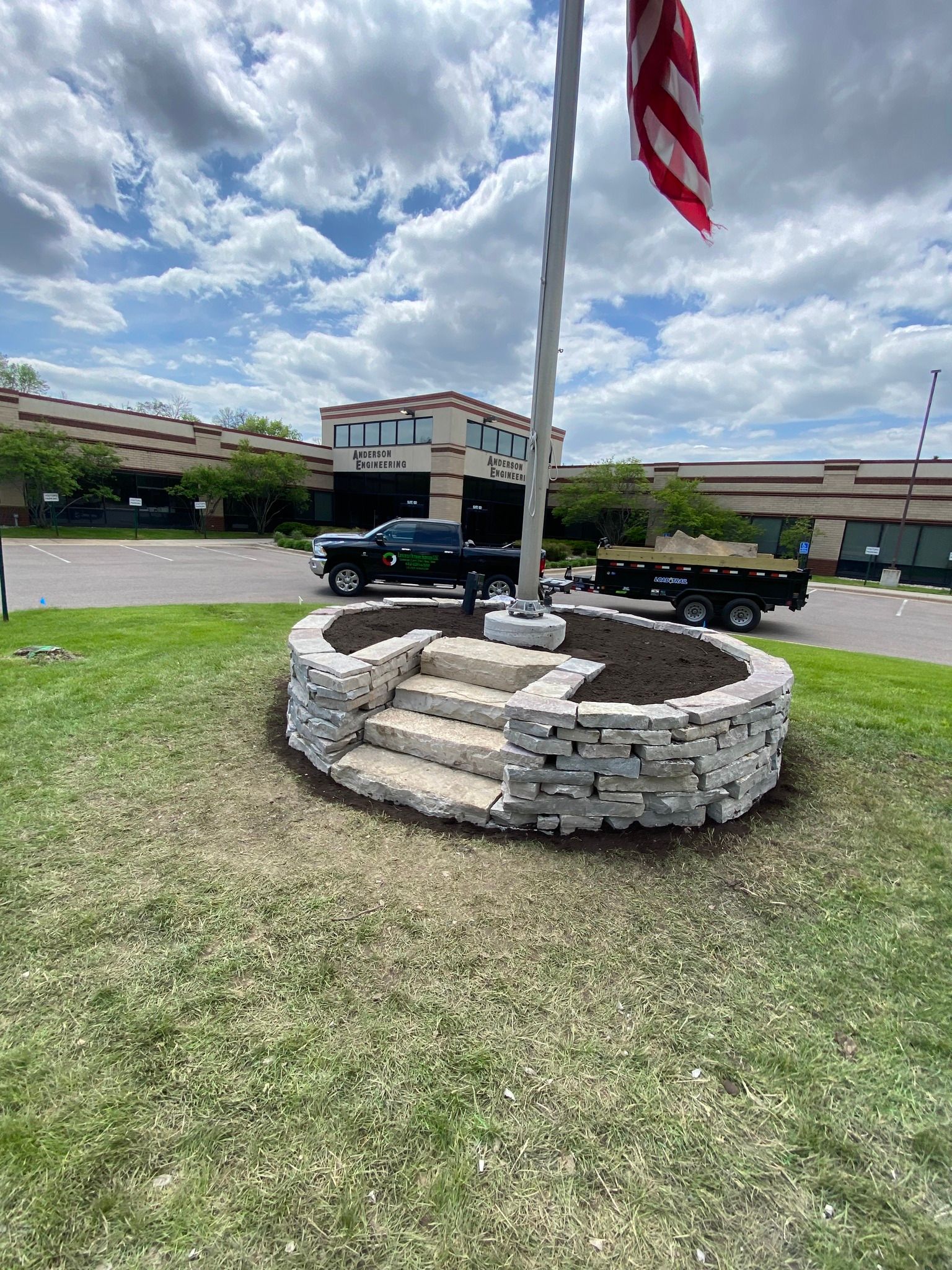 Flagpole with American flag, stone-walled flower bed, steps, grass, blue sky, parked truck.