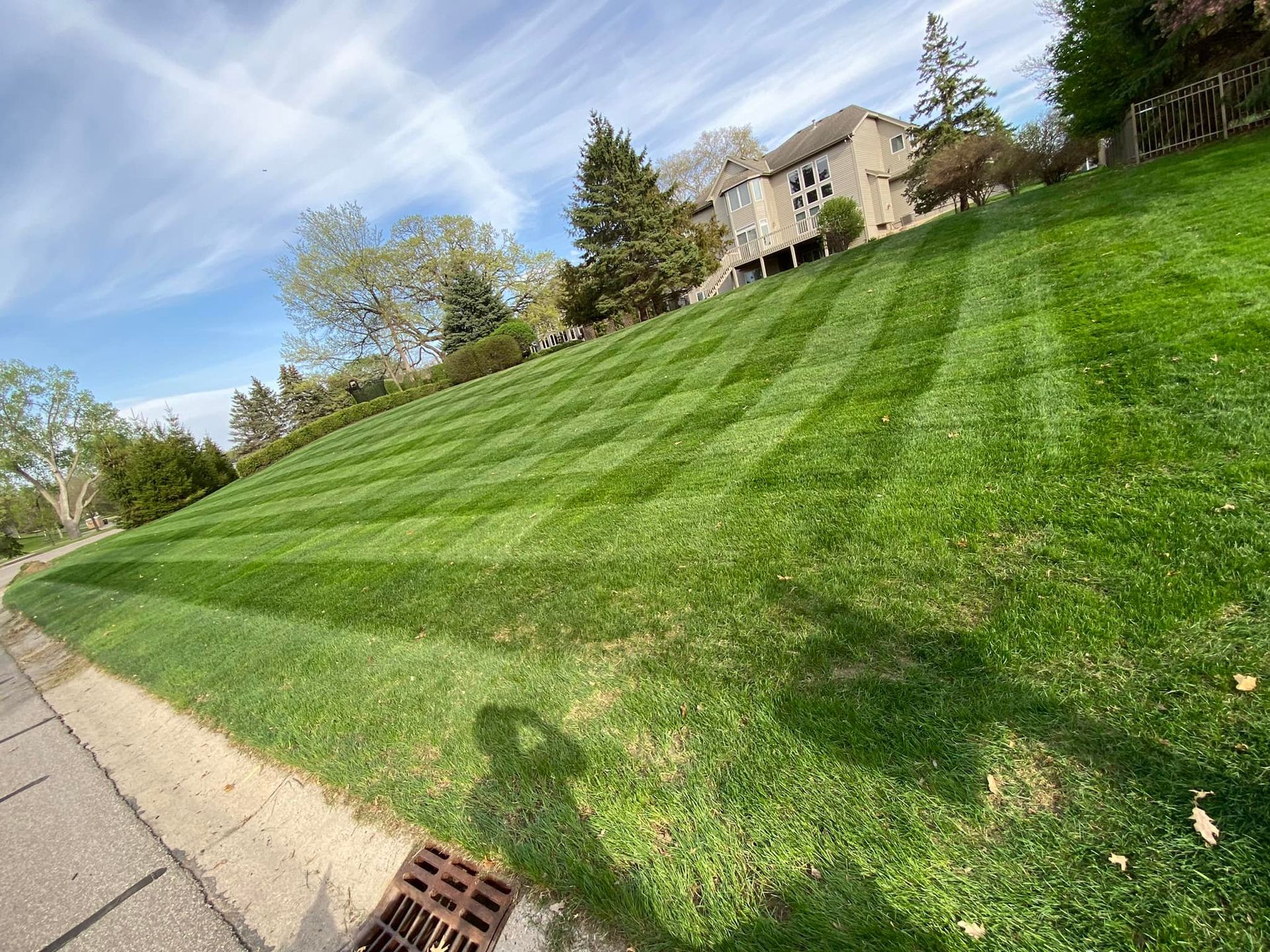 Lawn on a hillside, freshly mowed with striped patterns. Green grass, blue sky, and a house in the background.