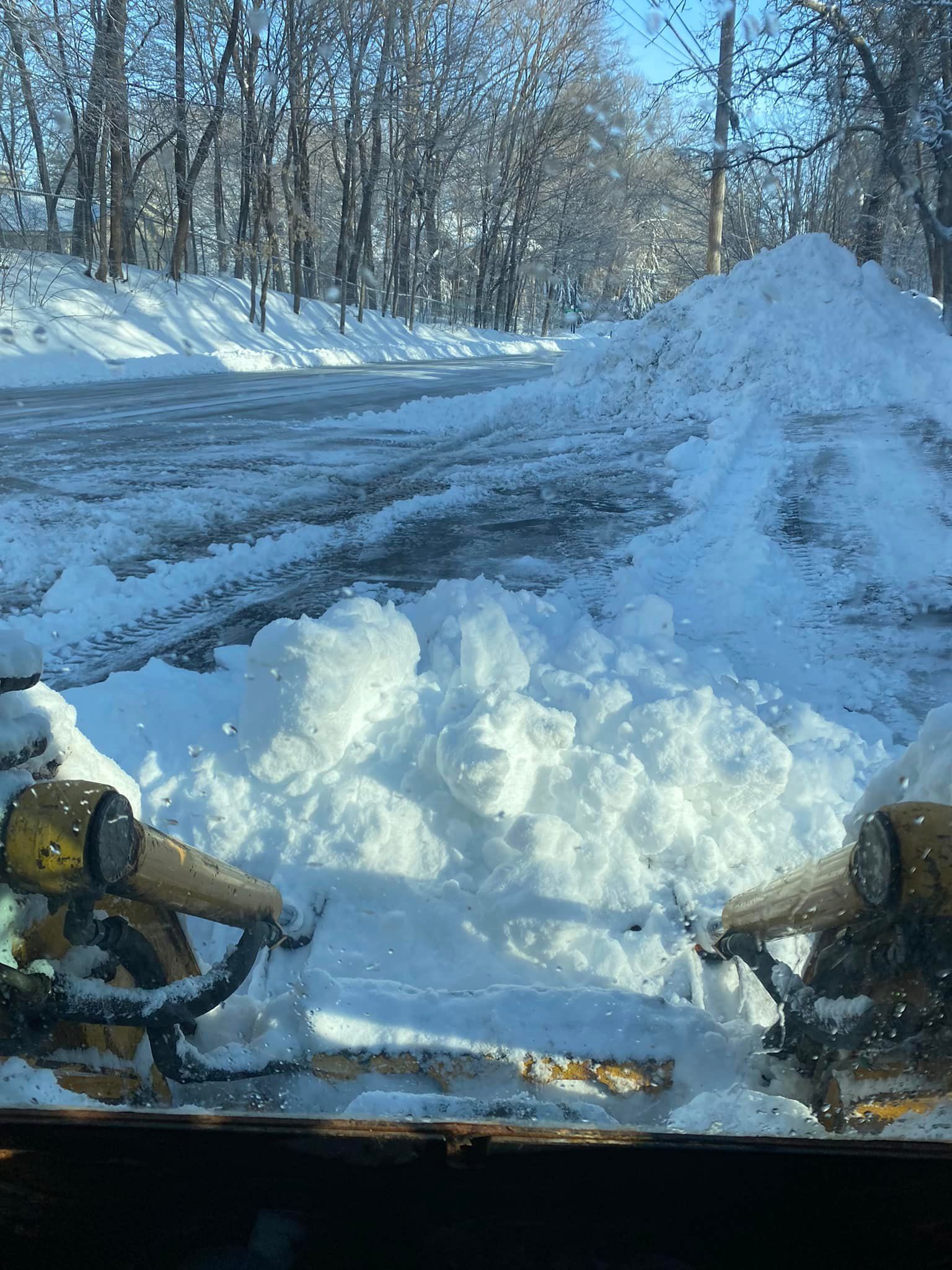 Snow plow clearing a snowy driveway. A large pile of snow is to the right. Trees are in the background.