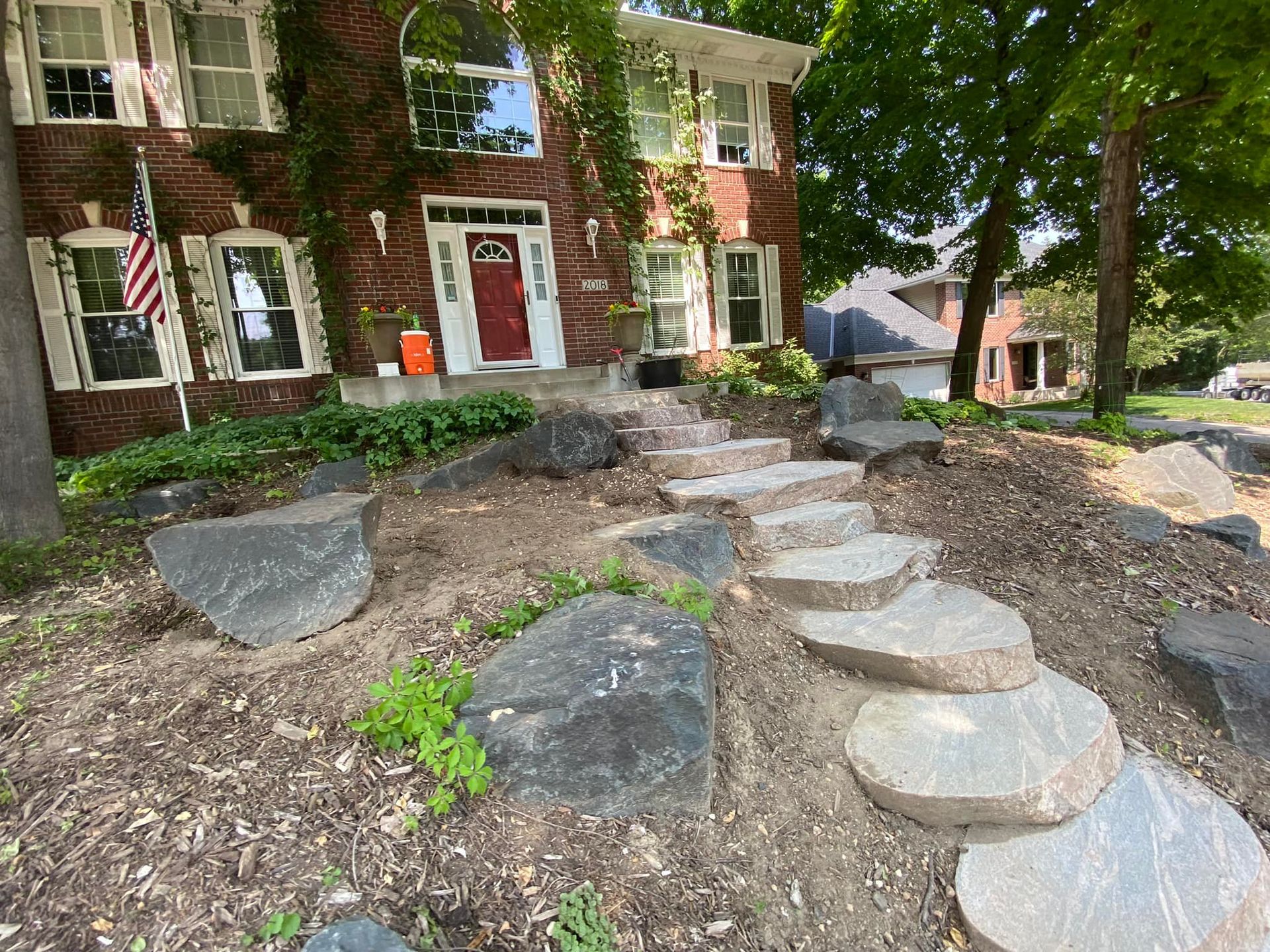 Stone steps leading up to a red-doored brick house with overgrown ivy. American flag on left.