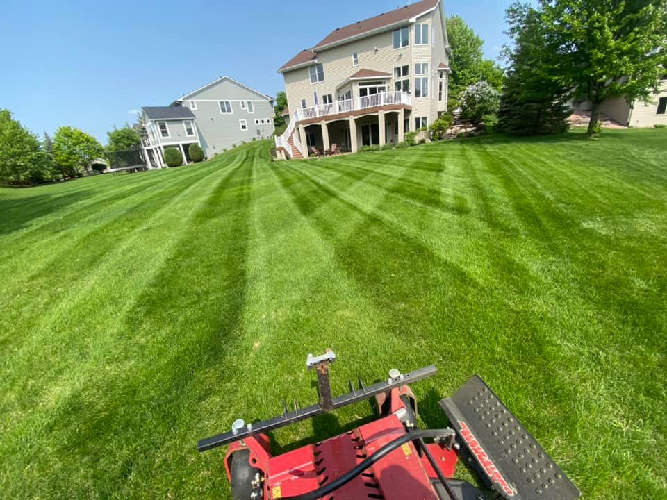 Lawn mower cutting stripes into a green grassy hill. Two houses in background under a blue sky.