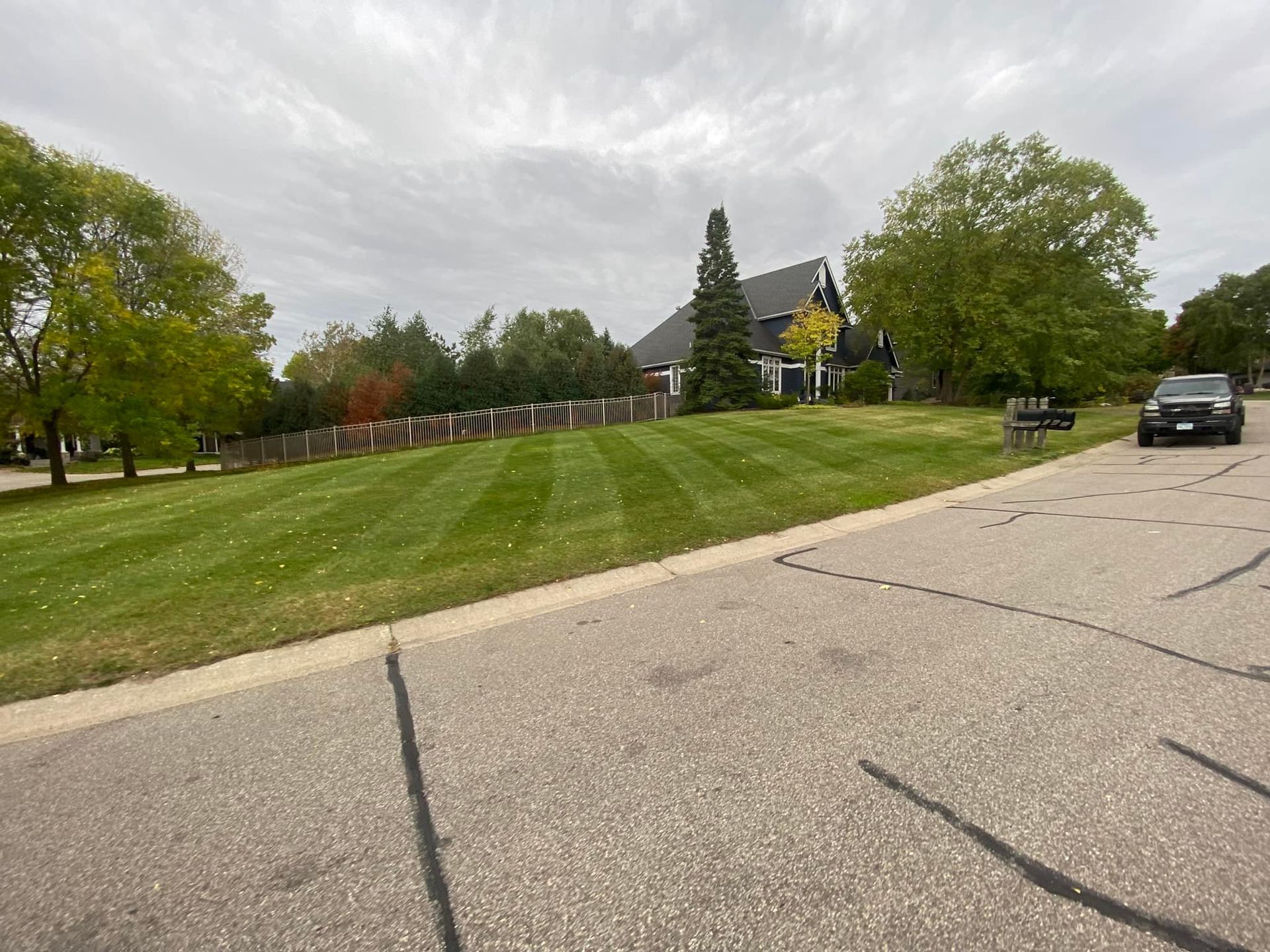 Lawn with mowed stripes, house with dark roof in background, car parked on road, cloudy sky.