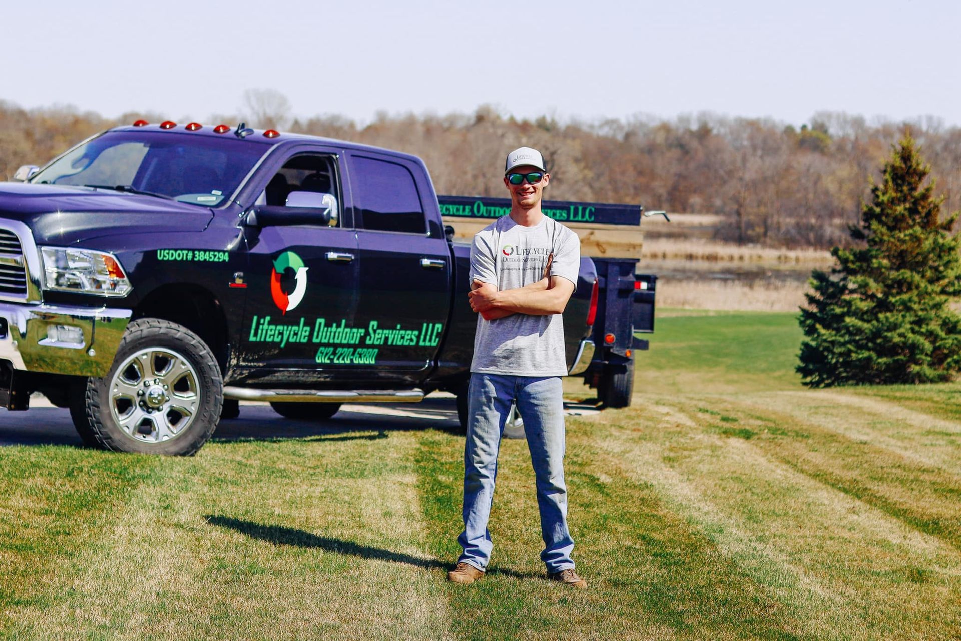 Man standing in front of a black pickup truck with landscaping service logo. Outdoor setting with a field and trees.