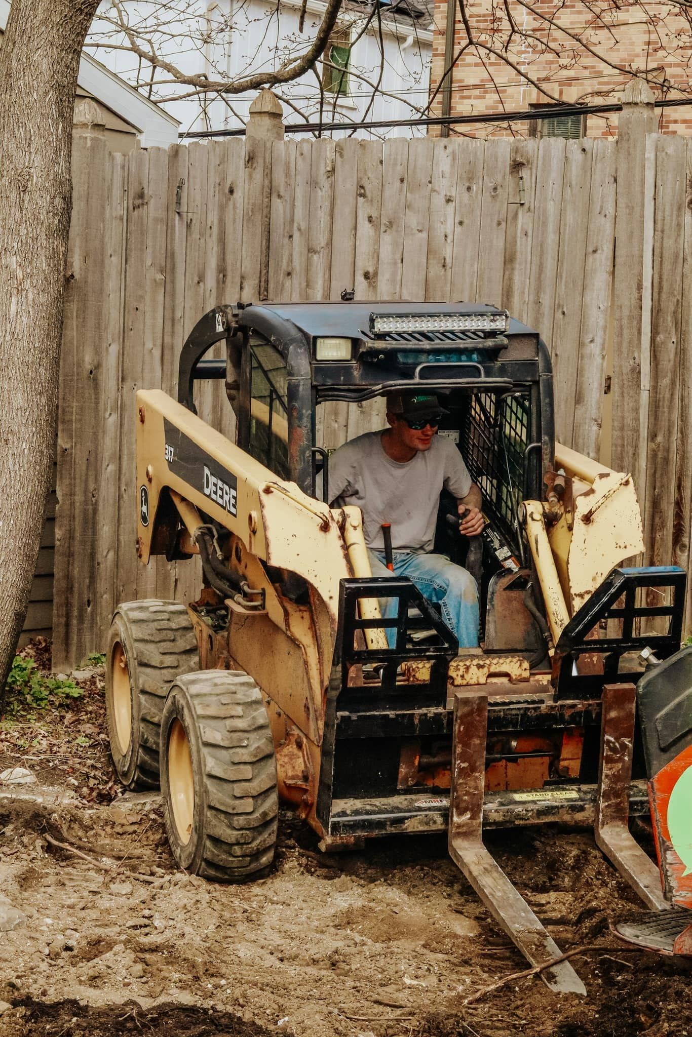 Yellow skid steer loader with a person inside working in a dirt area near a wooden fence.