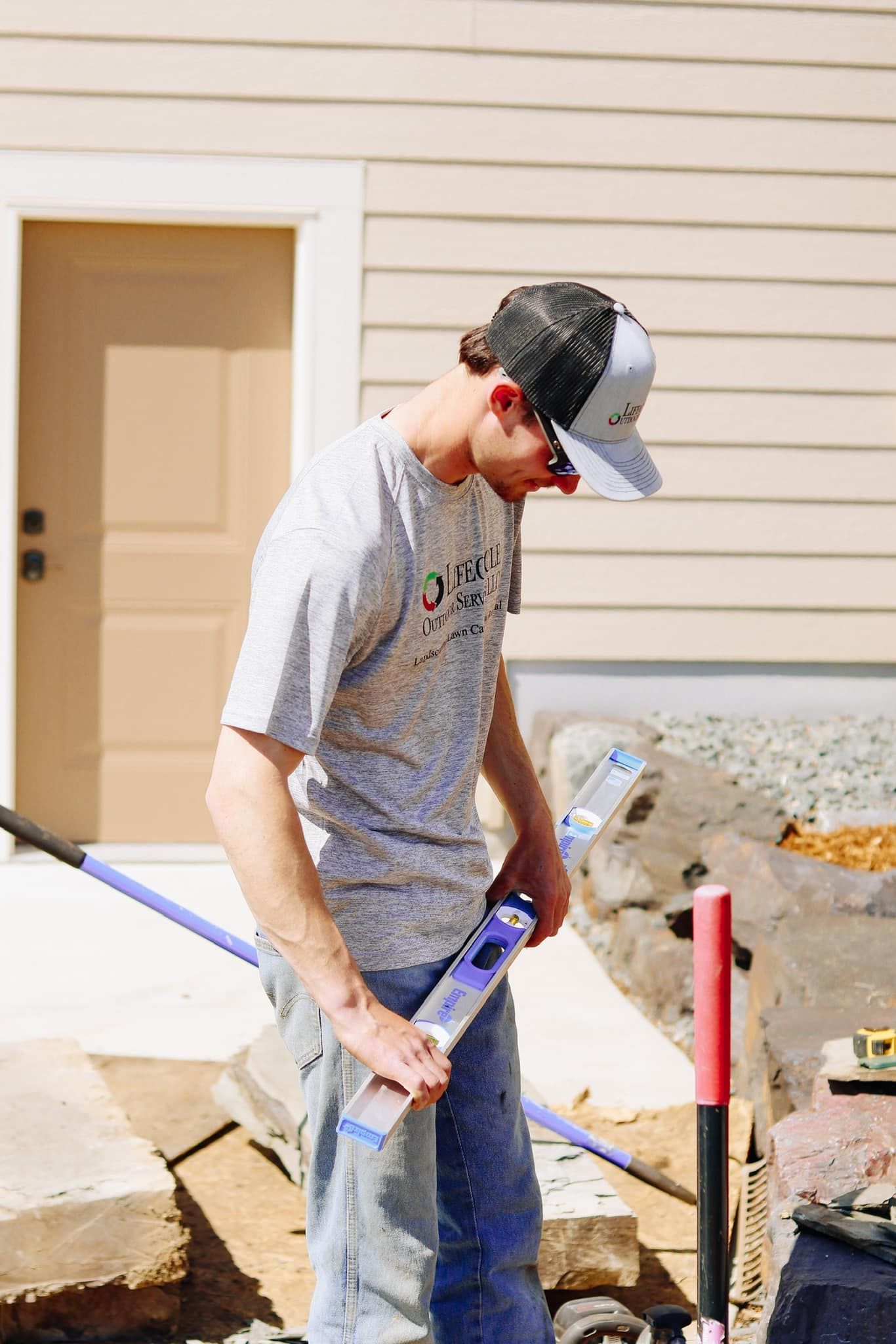Man in a baseball cap leveling stones with a tool outside a building.
