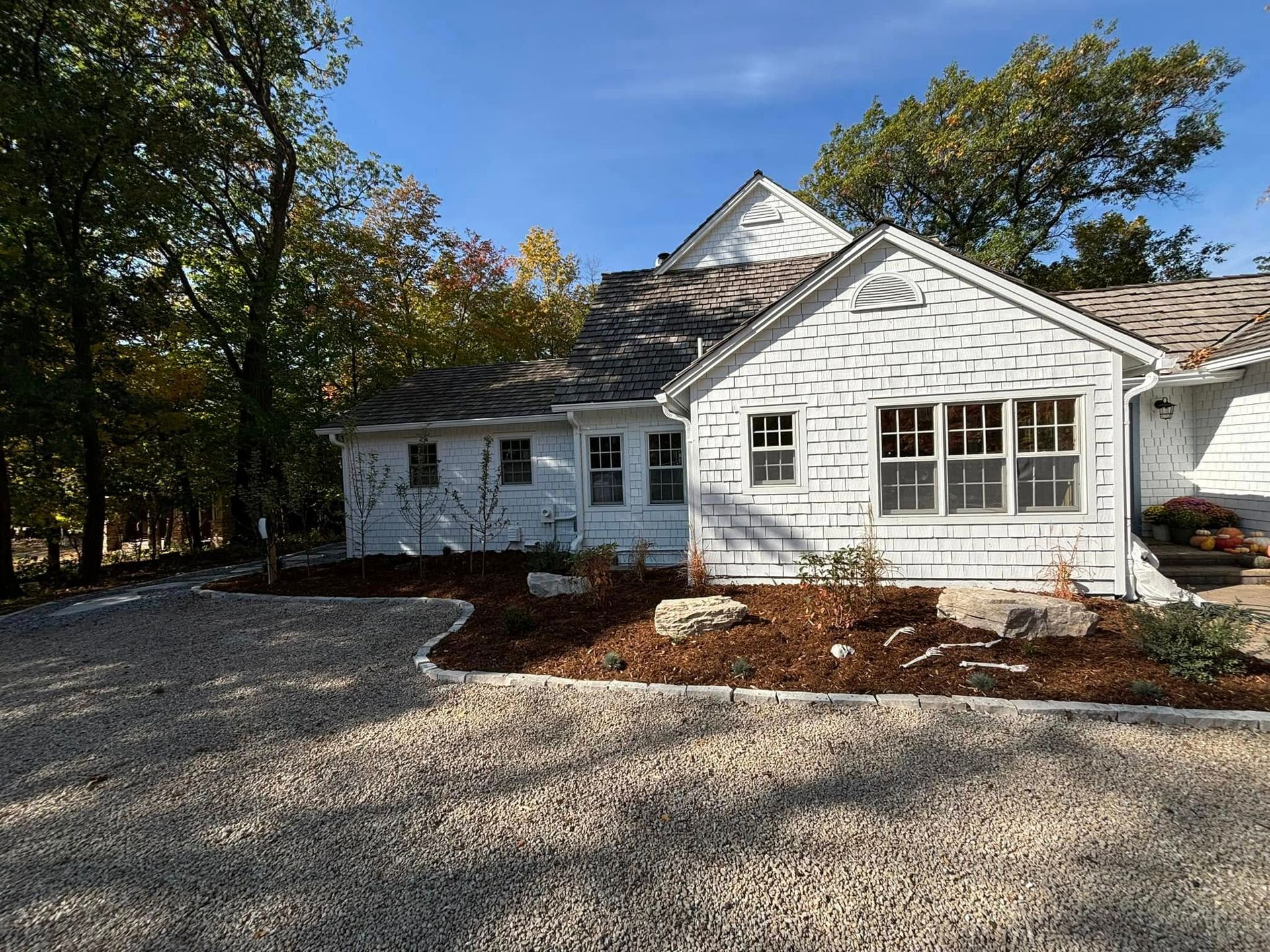 White house with weathered shingles, gravel driveway, and mulch landscaping under a blue sky.