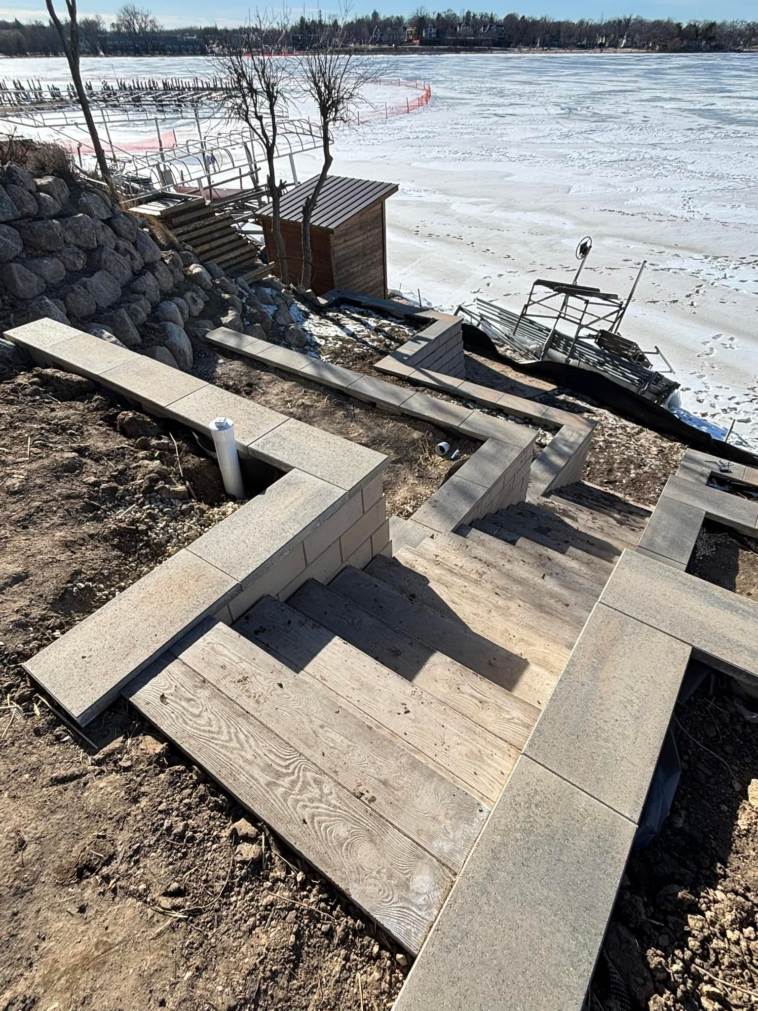 Stone staircase leading down to a frozen lake. Brown dirt surrounds the stairs and a small wooden shed sits at the top.