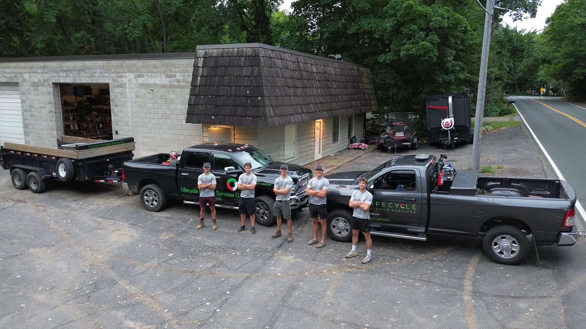 Five people pose with trucks and trailers outside a building, roadside.