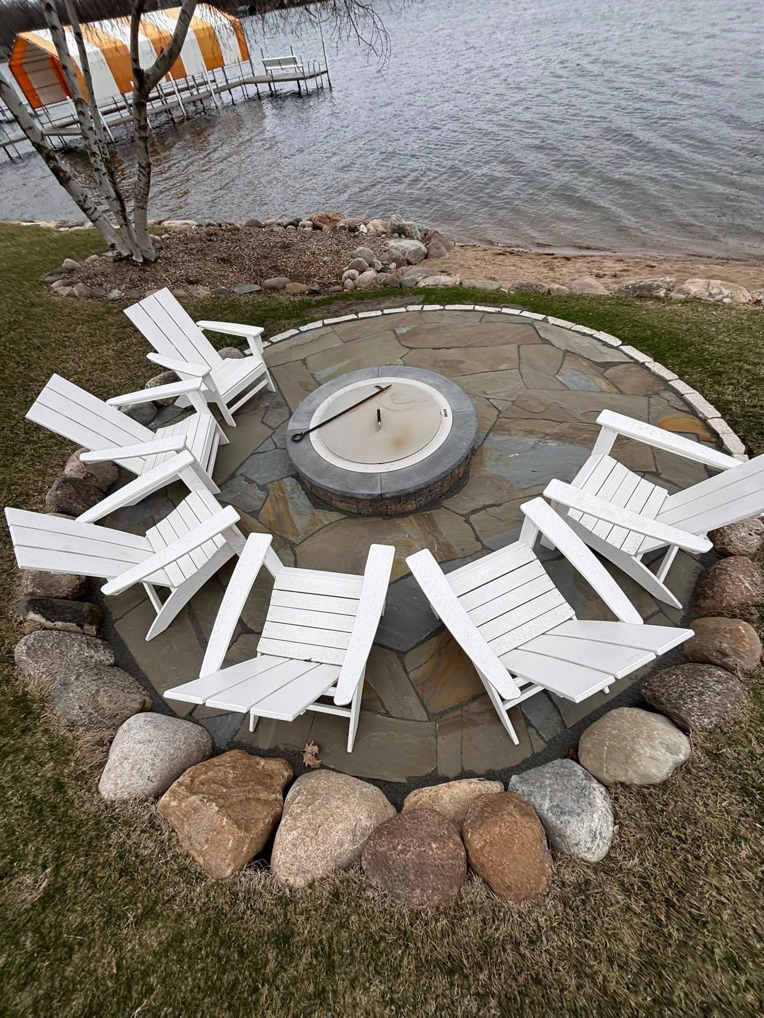A circular fire pit with white Adirondack chairs around it, near a lake.