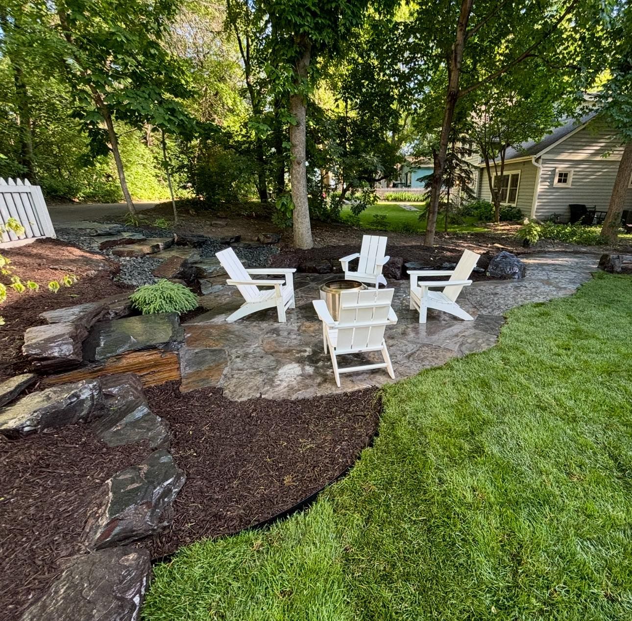 Outdoor patio with white chairs on a stone surface, surrounded by mulch, rocks, and green grass.