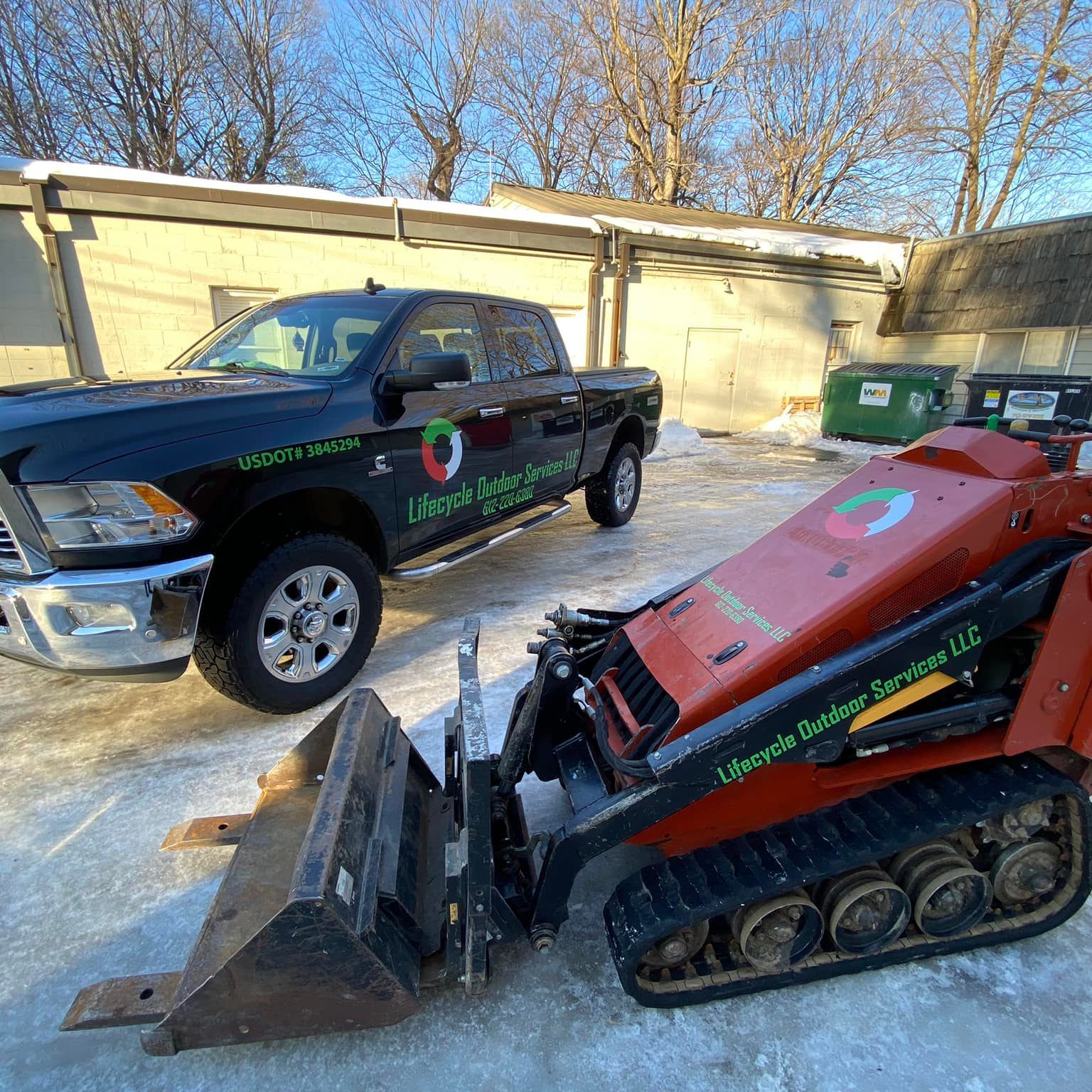 Black pickup truck and red mini-excavator with company logos, parked in front of a building on a sunny day.