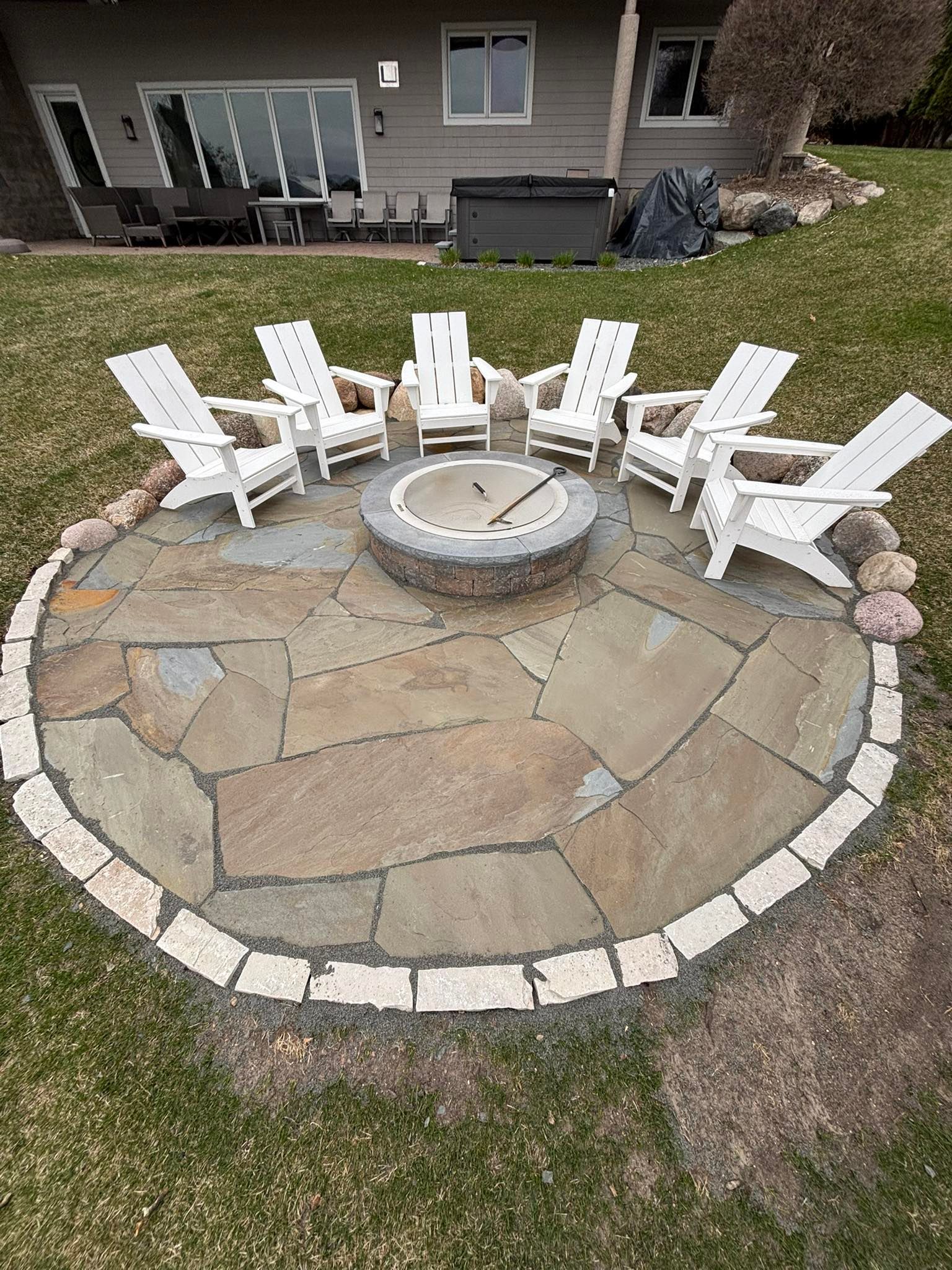 Fire pit area with six white Adirondack chairs on a stone patio, surrounded by grass.