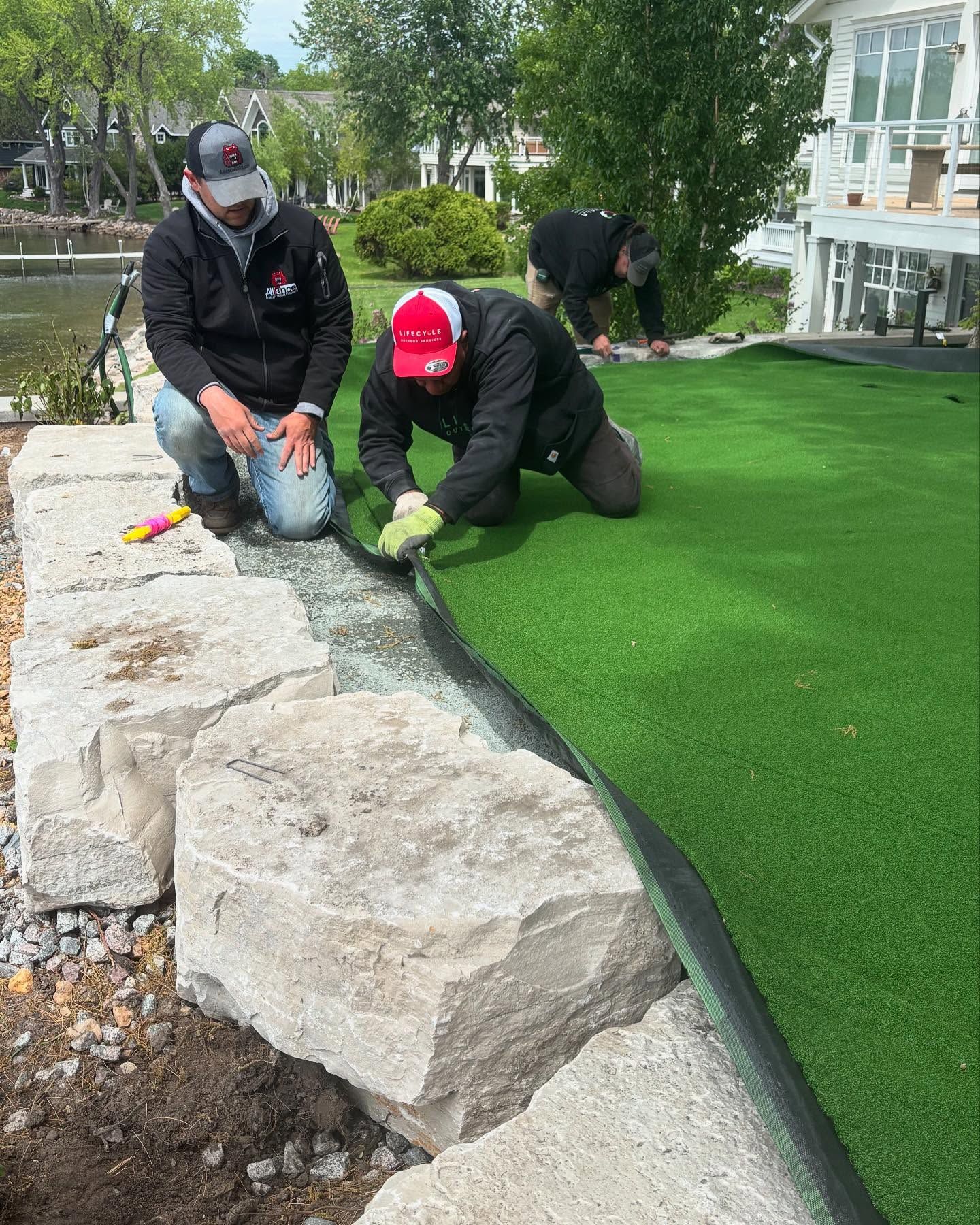 Men installing green artificial turf next to a stone wall; one kneels, two stand nearby, outdoors.
