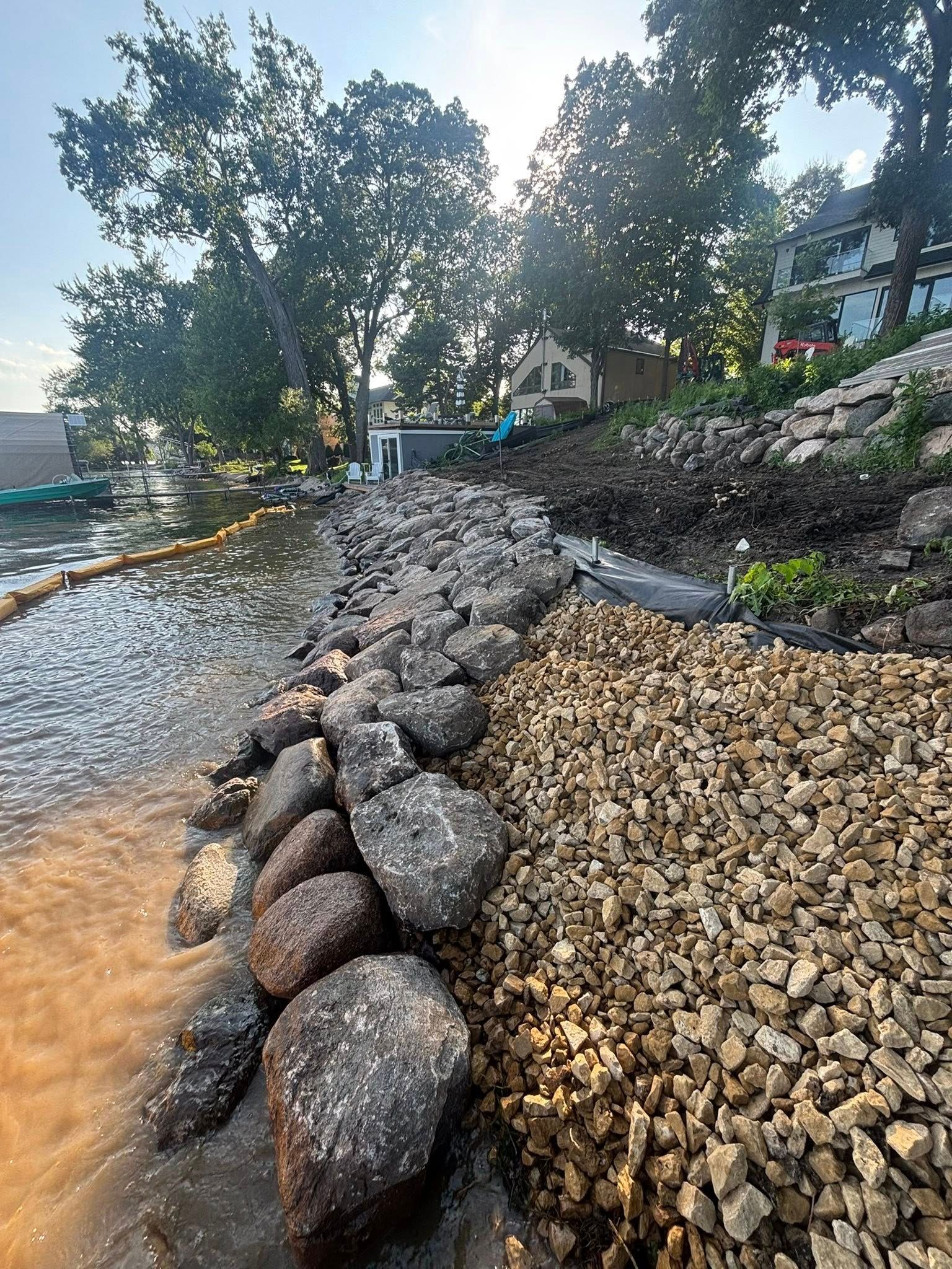 Lakeshore erosion control project: large rocks and gravel alongside a shoreline, trees and houses in the background.