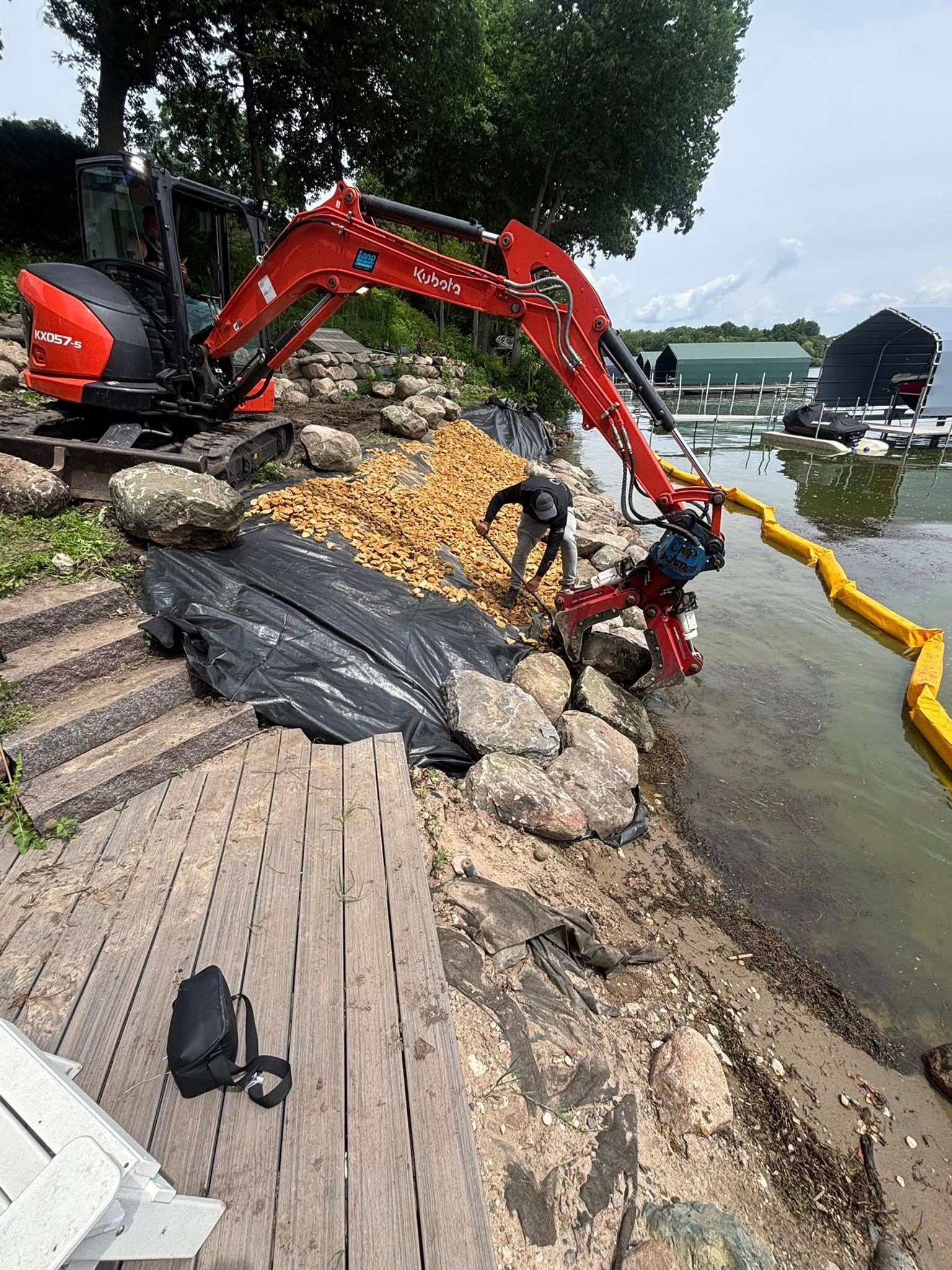 Excavator and workers on a rocky shoreline, removing debris near a lake.