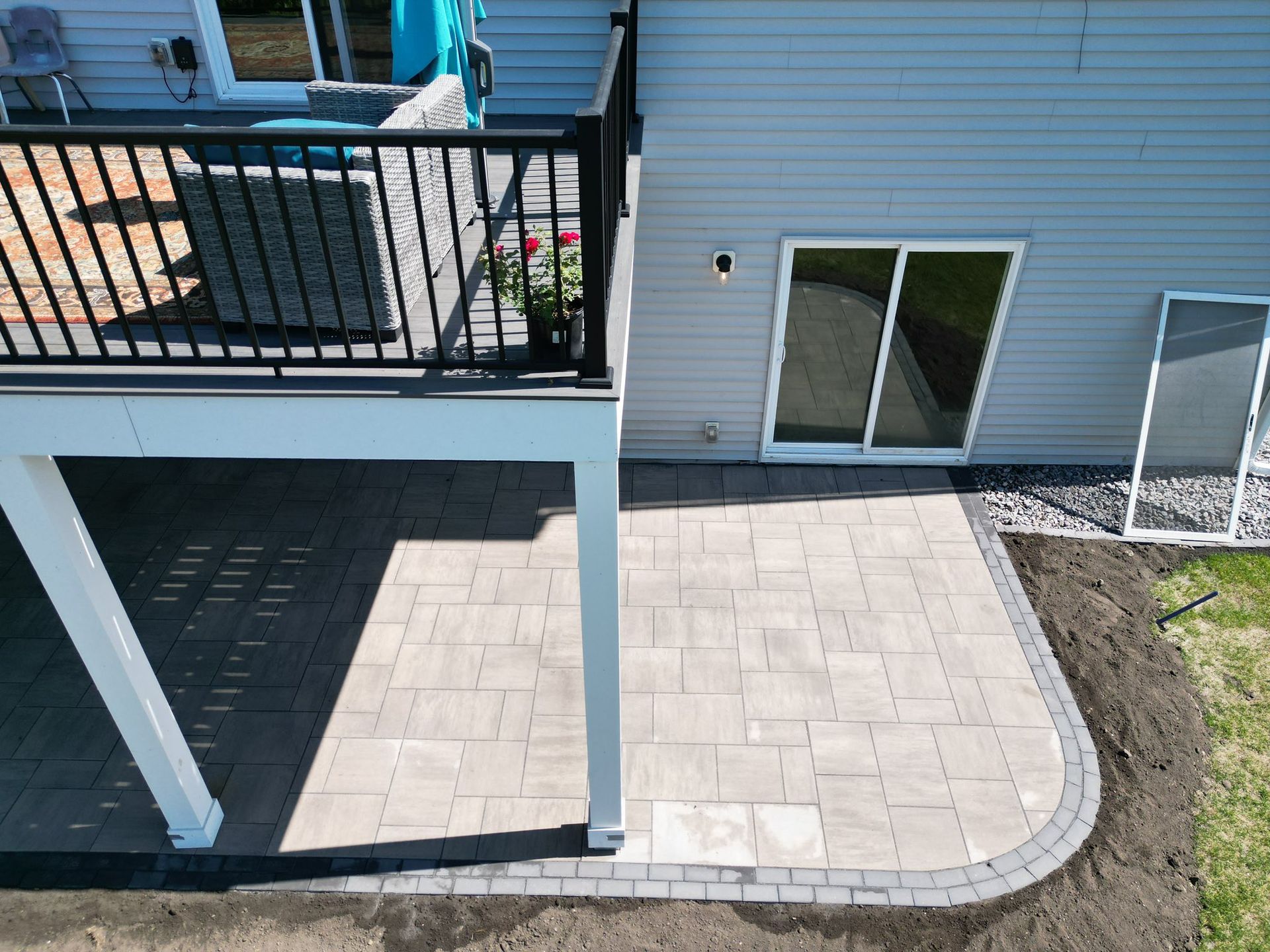 Paver patio beneath a deck, gray pavers with a black border, next to a sliding glass door and house.