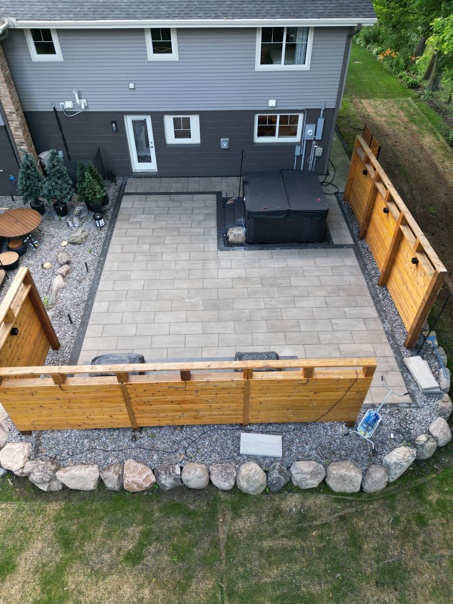 Backyard patio with hot tub, bordered by wooden fences and stone. Grey house in the background, green lawn.