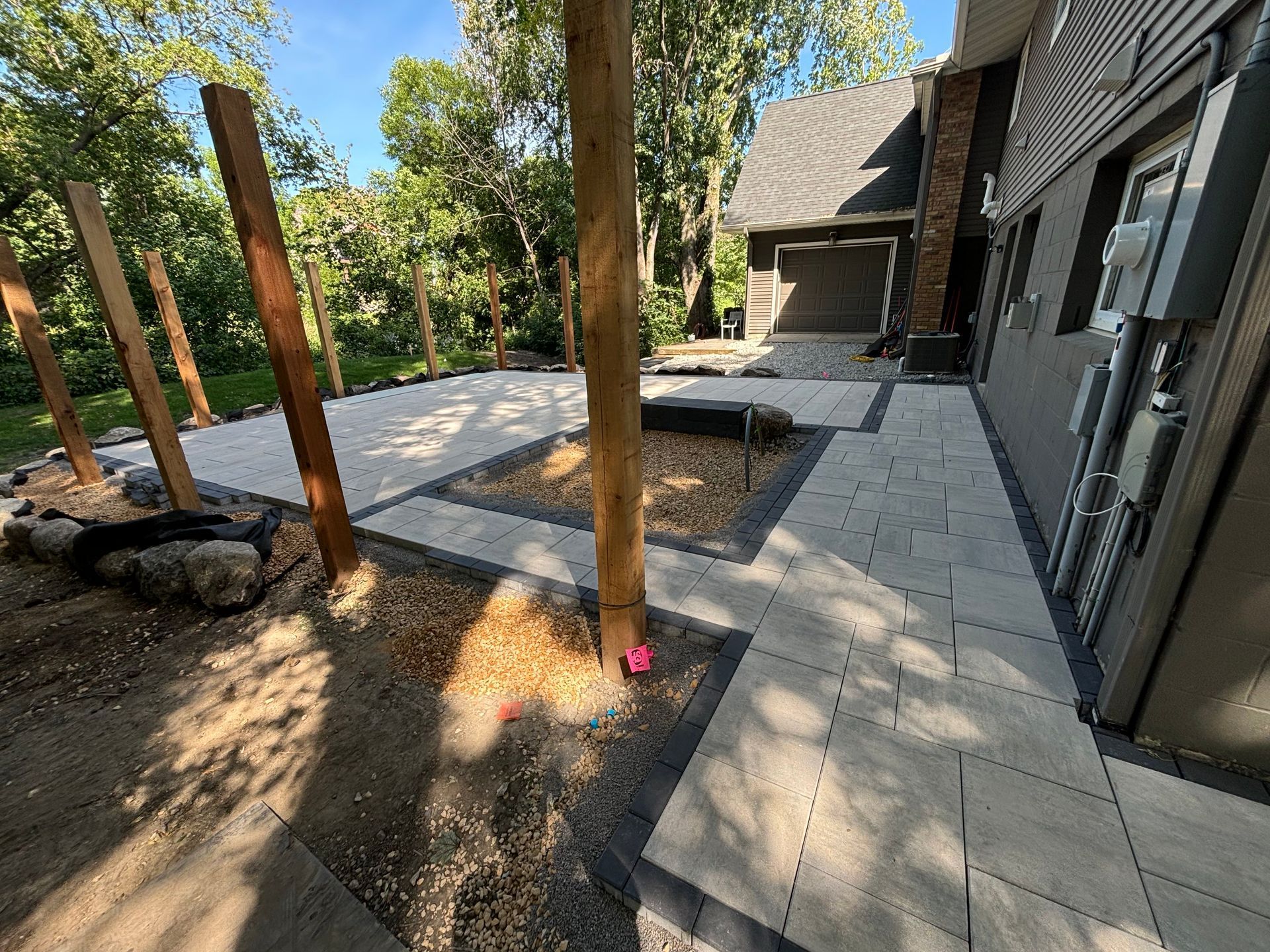 Patio with pavers and gravel beds, trees, and wooden posts next to a modern building with garage.