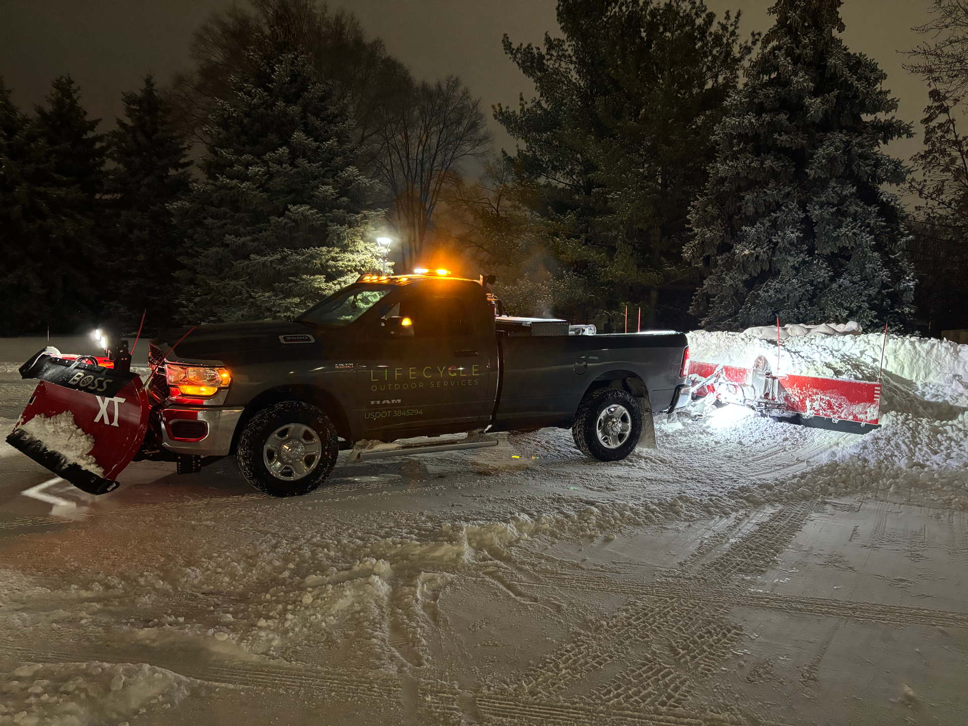 A truck with a snowplow clearing snow in a snowy outdoor area at night. Orange lights are on.