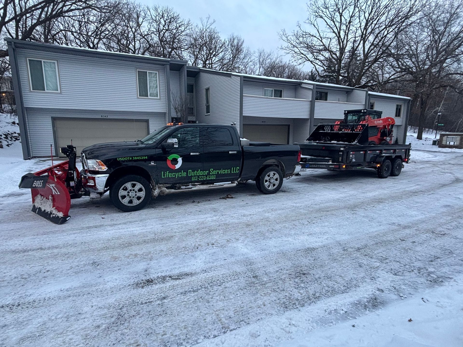 Black pickup truck with snowplow towing a trailer with landscaping equipment in front of a building covered in snow.