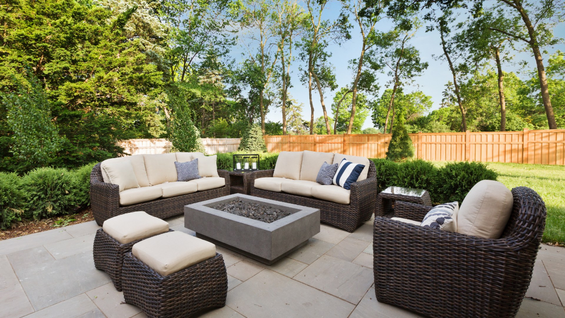 Patio with wicker furniture, fire pit, and stone pavers in a backyard with trees and a wooden fence.