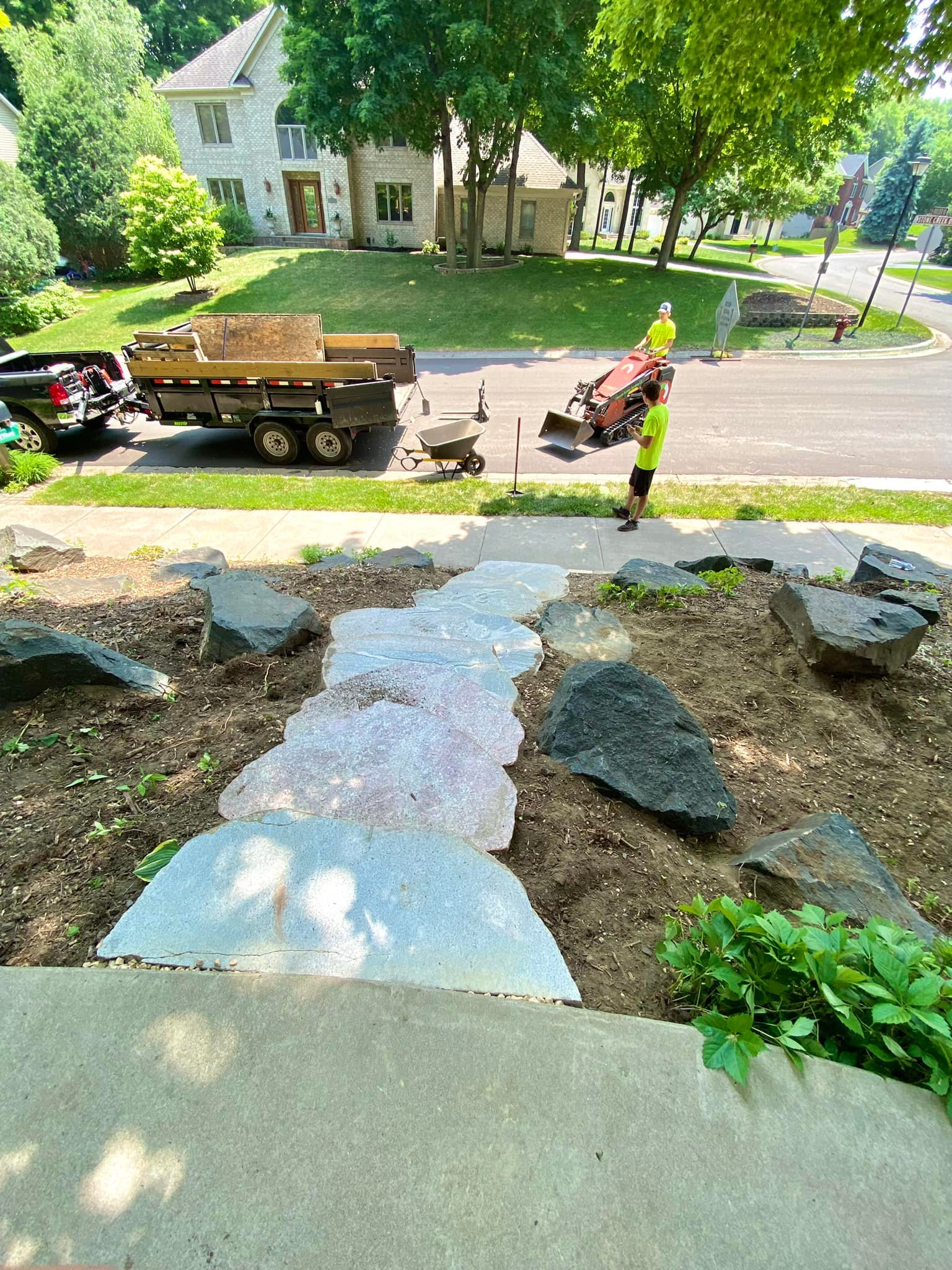 Workers install a stone walkway; truck and trailer with supplies in the background.