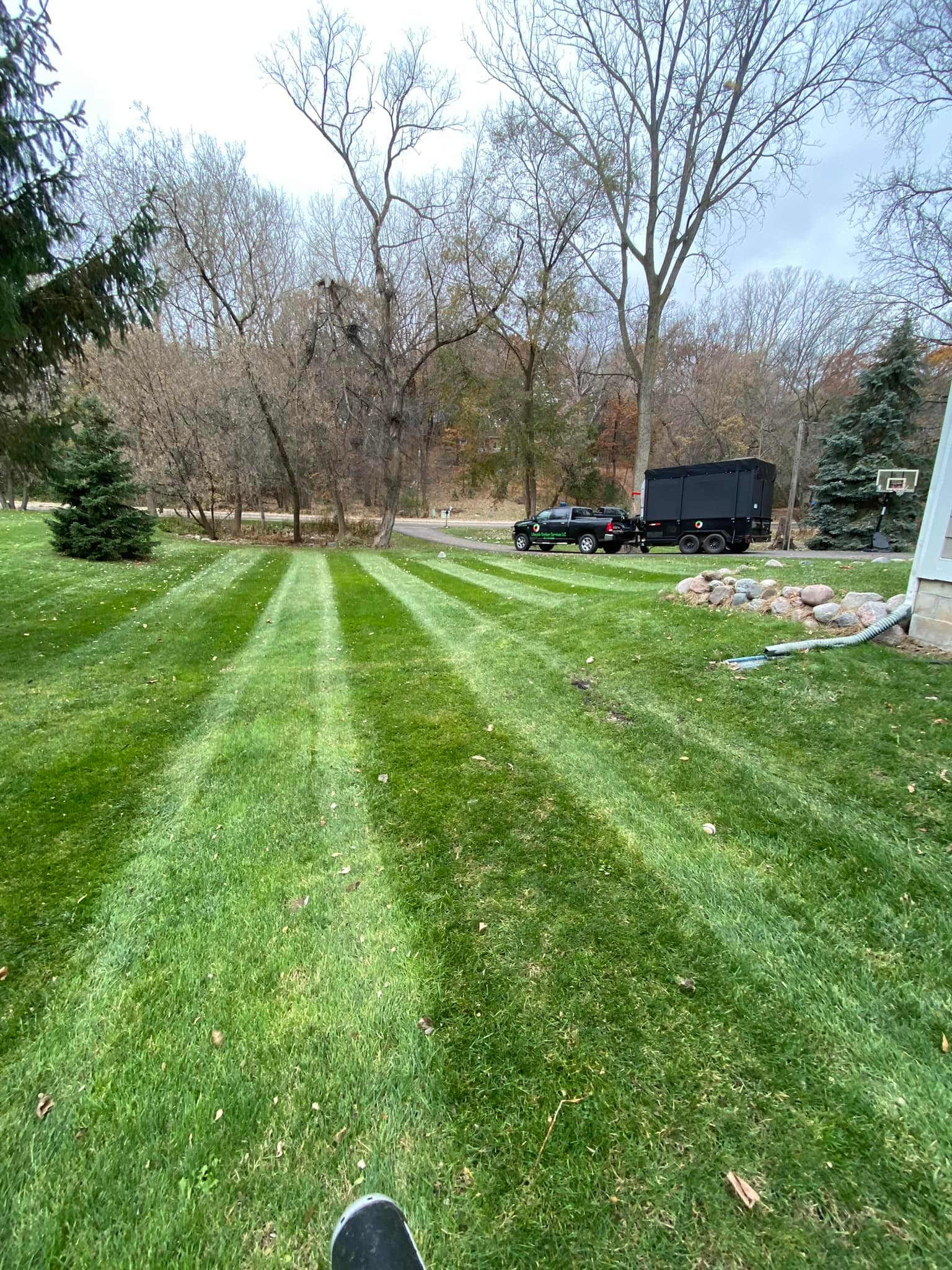 Lawn with striped pattern, black truck with trailer in the background, trees, and cloudy sky.
