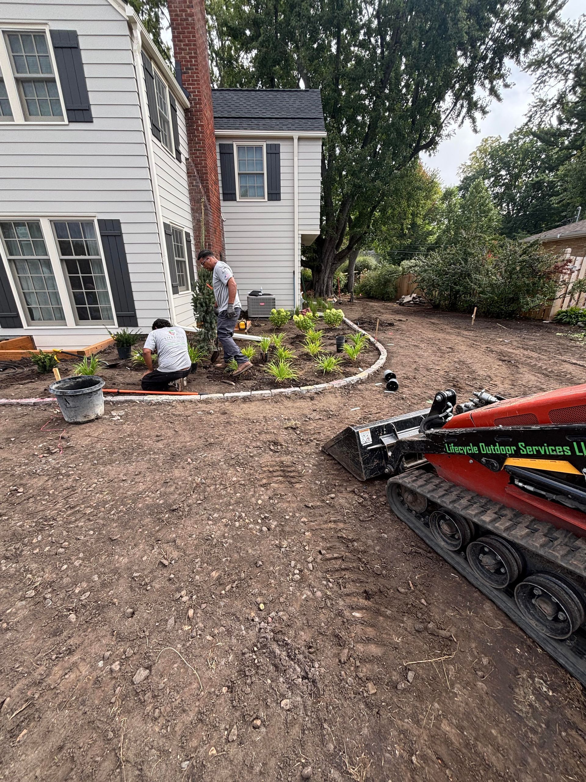 Two workers landscaping a yard next to a two-story white house with black shutters, using a small earth-moving machine.