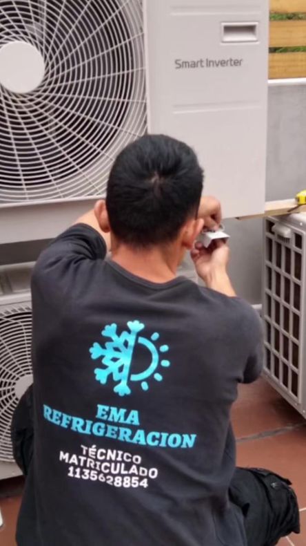 Técnico con camisa negra reparando una unidad de aire acondicionado exterior junto a la pared de un edificio.
