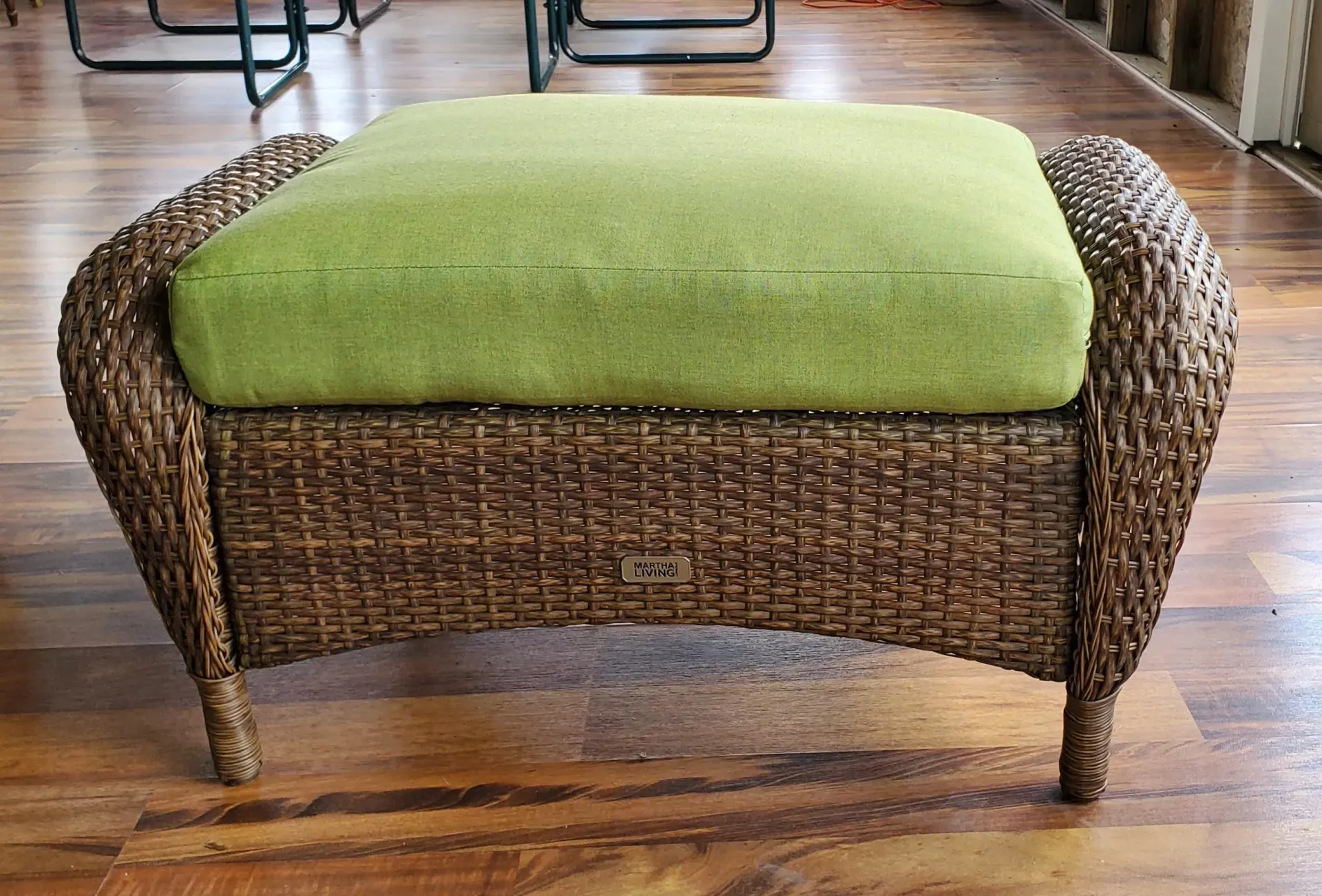 A wicker ottoman with a green cushion on a wooden floor