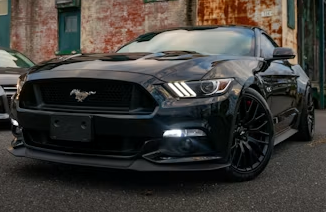 Black Ford Mustang parked on a street, front view with headlights on and weathered brick wall behind it