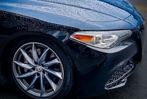 Close-up of a dark blue sports car’s front wheel, headlight, and grille on a wet road