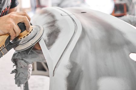 Person sanding a car panel with an electric sander in a garage; panel is primed white.