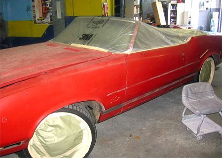 Red convertible car in a garage, prepped for painting. Wheels and interior covered in protective material.