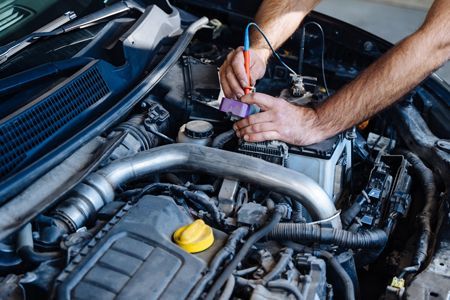 Mechanic's hands testing a car's engine with a multimeter; battery, hoses, and tubes visible.