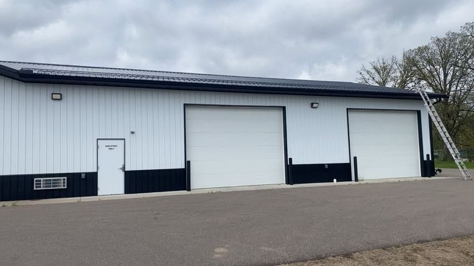 White industrial warehouse with a large roll-up door, side entry door, and ladder under an overcast sky